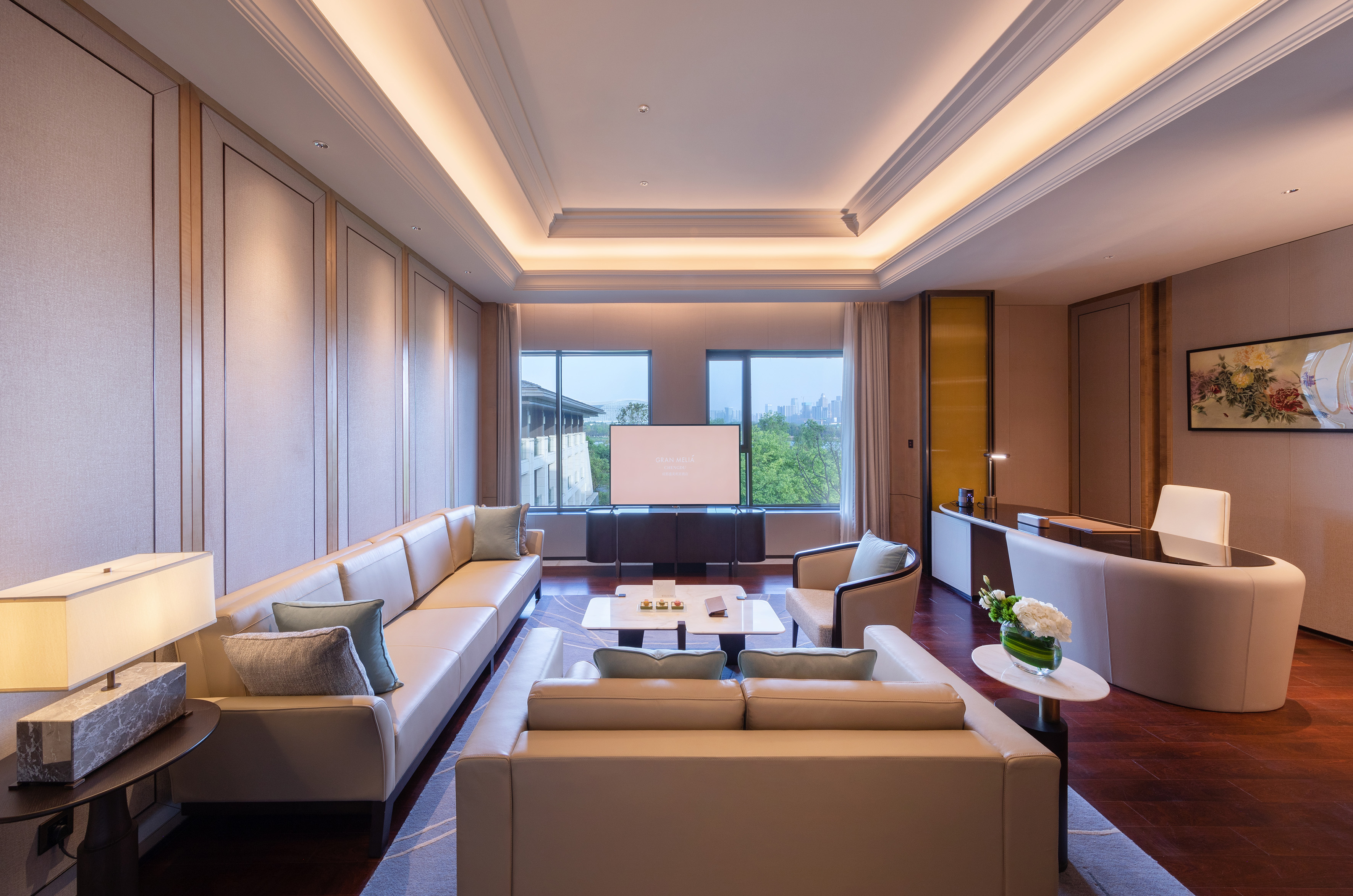 Spacious luxury hotel suite living room with sofa, TV, desk, and city view.