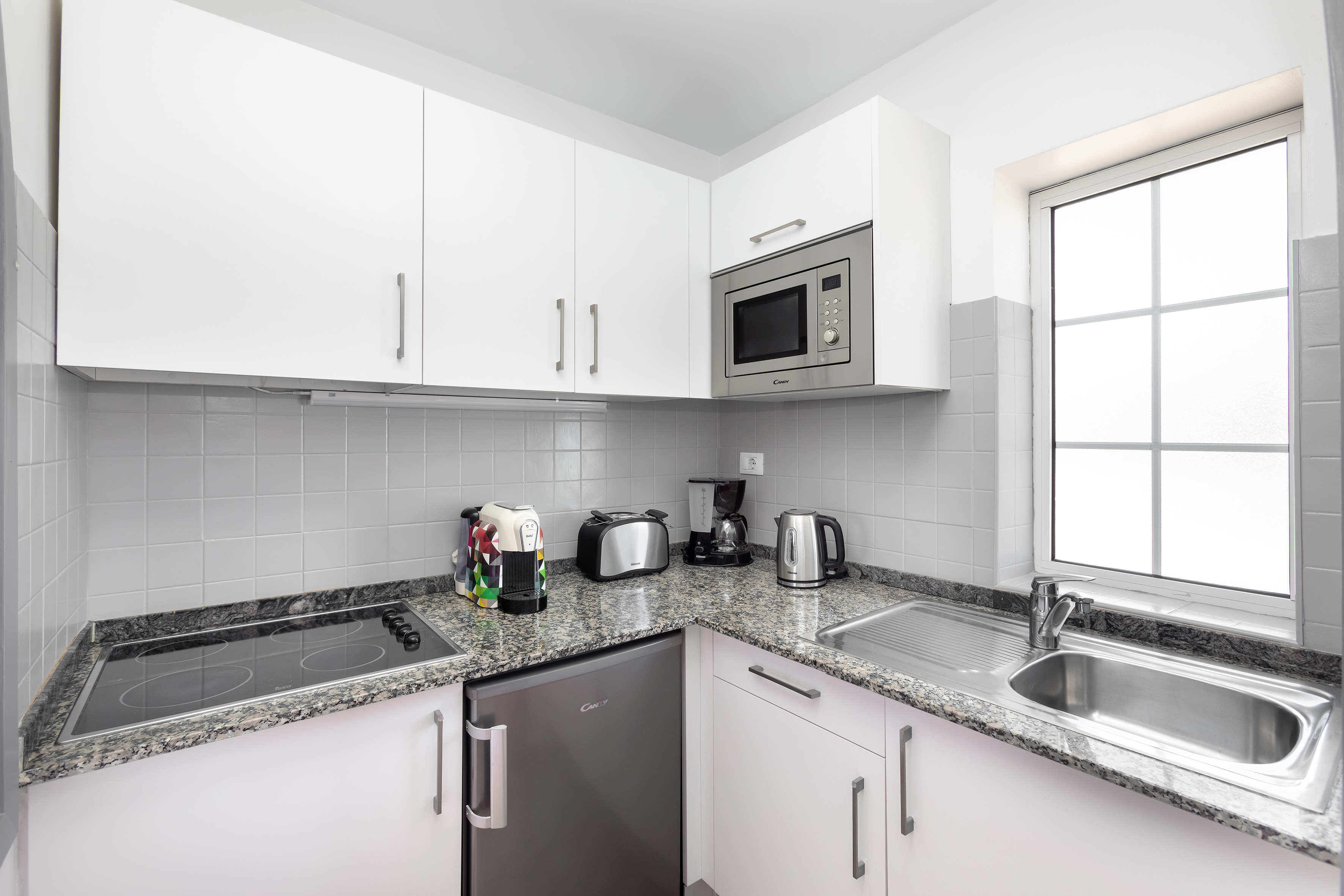 Modern white kitchen with granite countertops, microwave, hob, sink, and fridge.