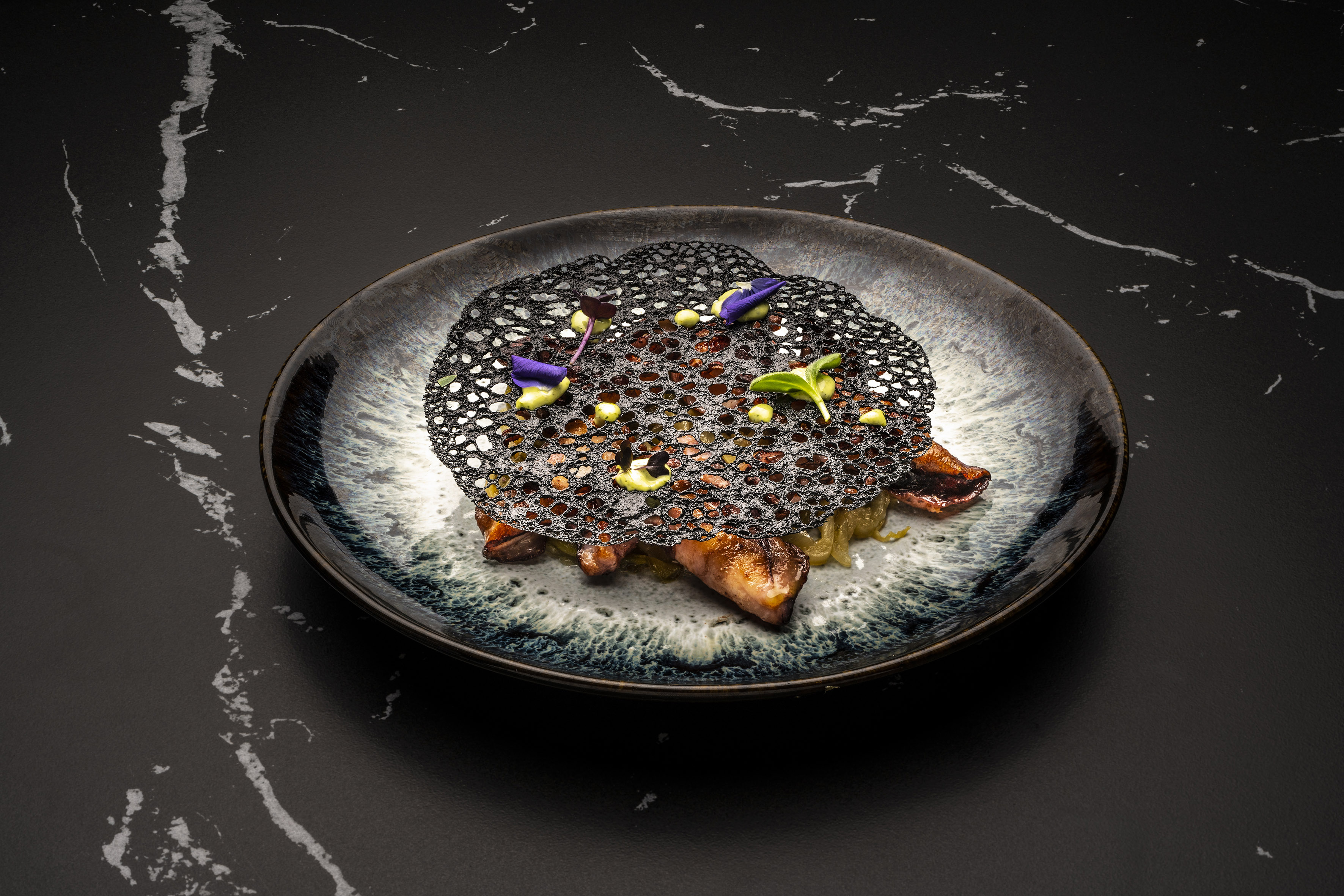 Modern plated dish: black lace tuile, seared meat, purple flowers, green garnish.