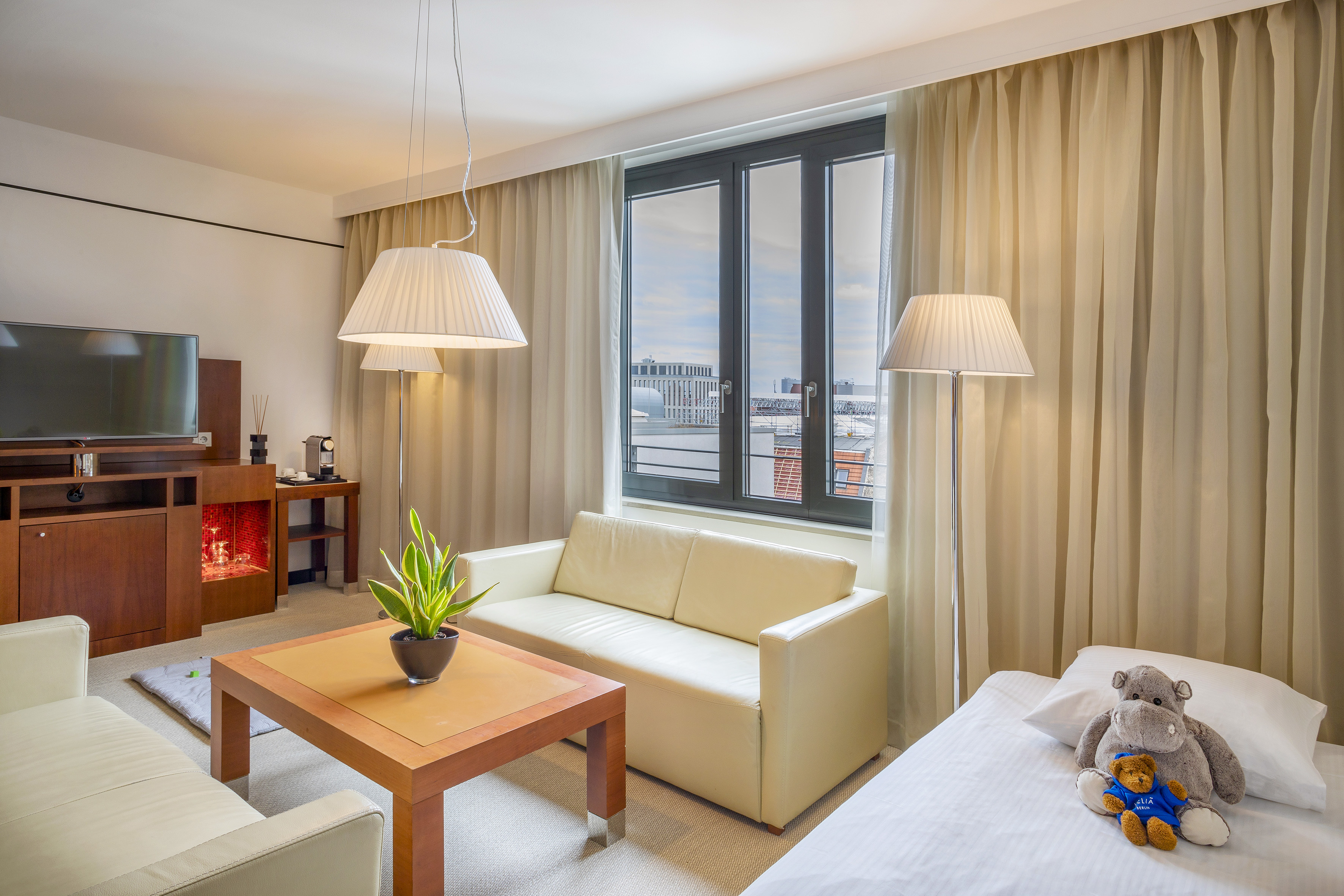 Hotel suite living area with sofa, television, single bed, and city view window.