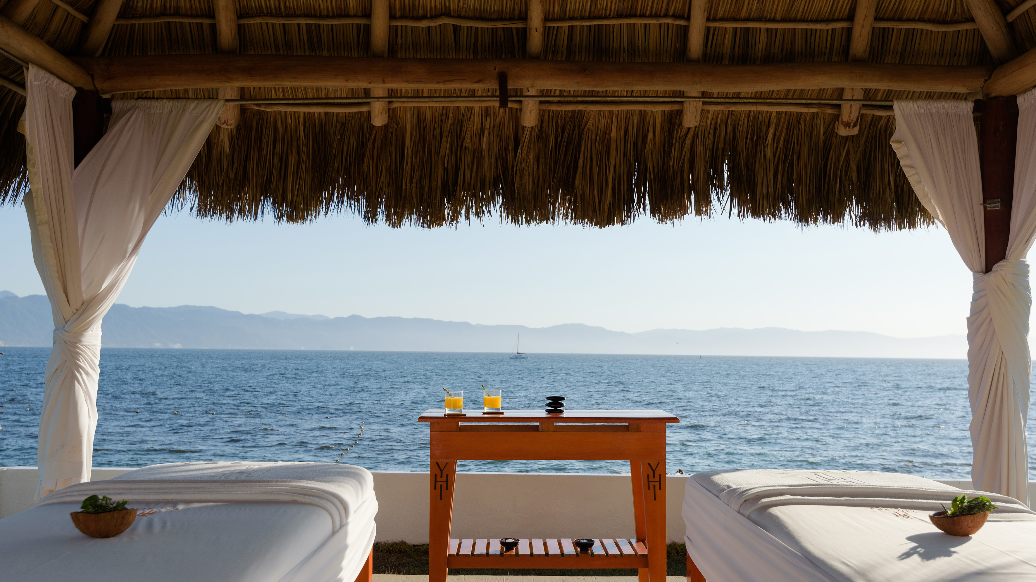 Tropical outdoor spa with ocean view, massage beds, palapa, and drinks.