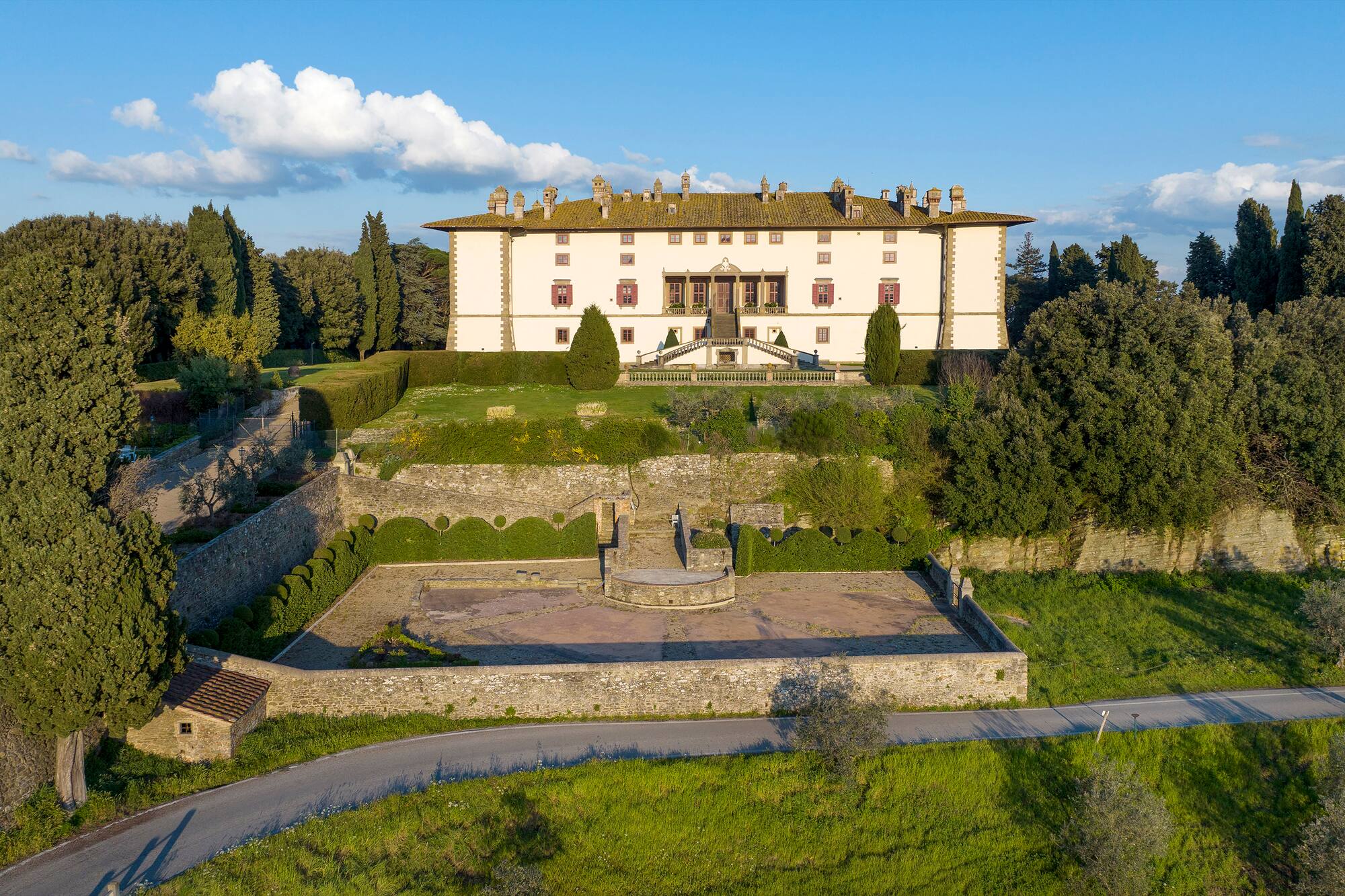 Large white Italian villa on hill, terraced gardens, cypress trees, winding road.