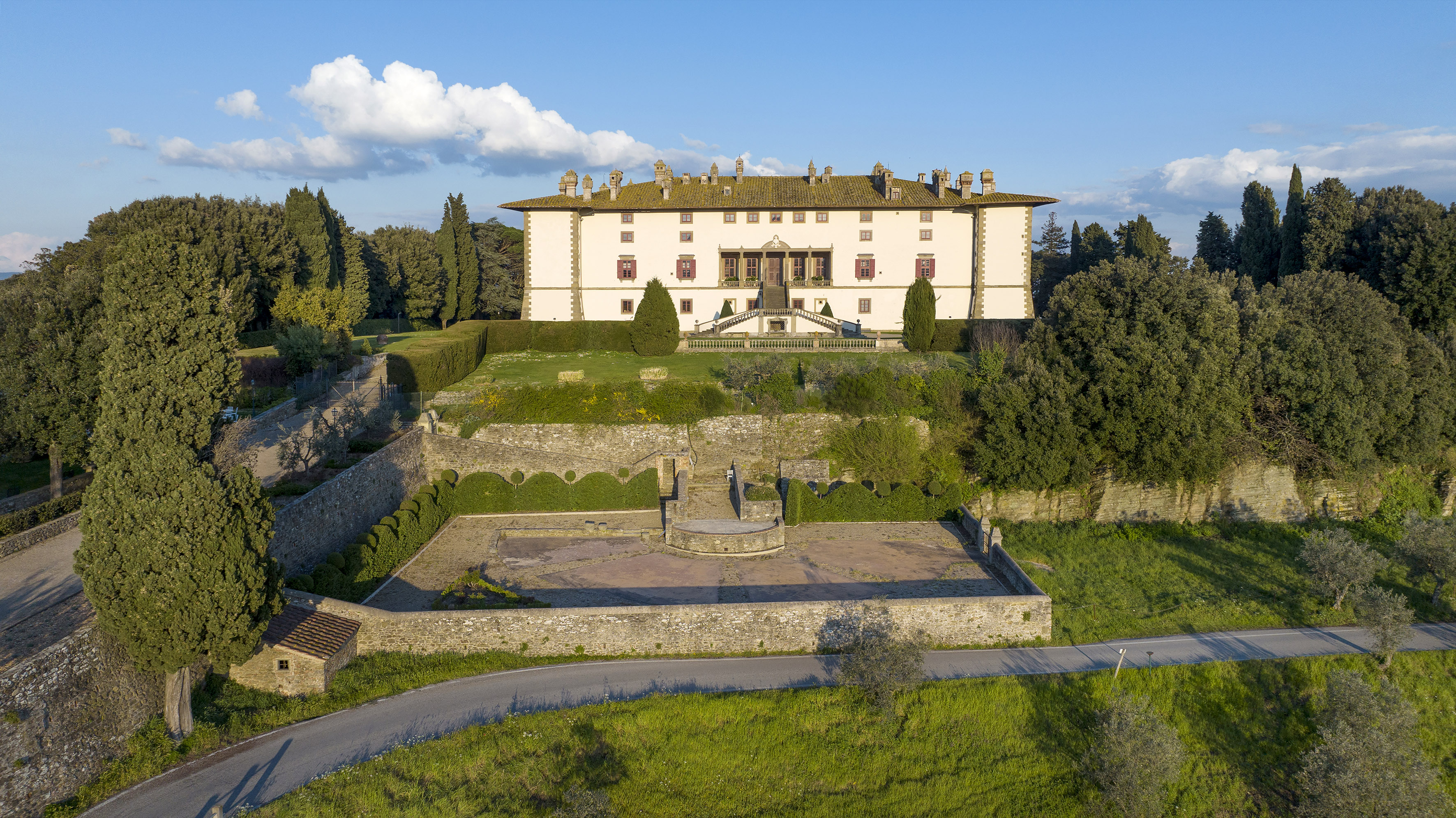 Large white Italian villa on hill, terraced gardens, cypress trees, winding road.