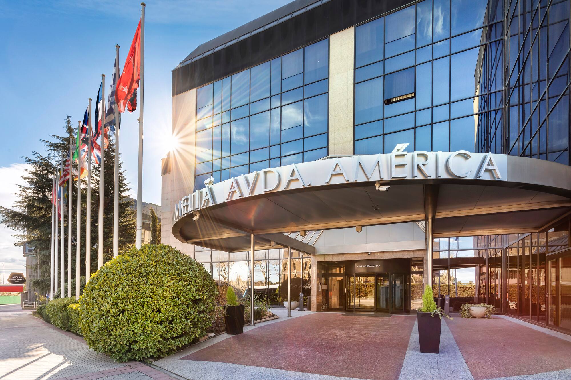 Melia Avda. America hotel entrance, modern glass building, flags, bright sun.