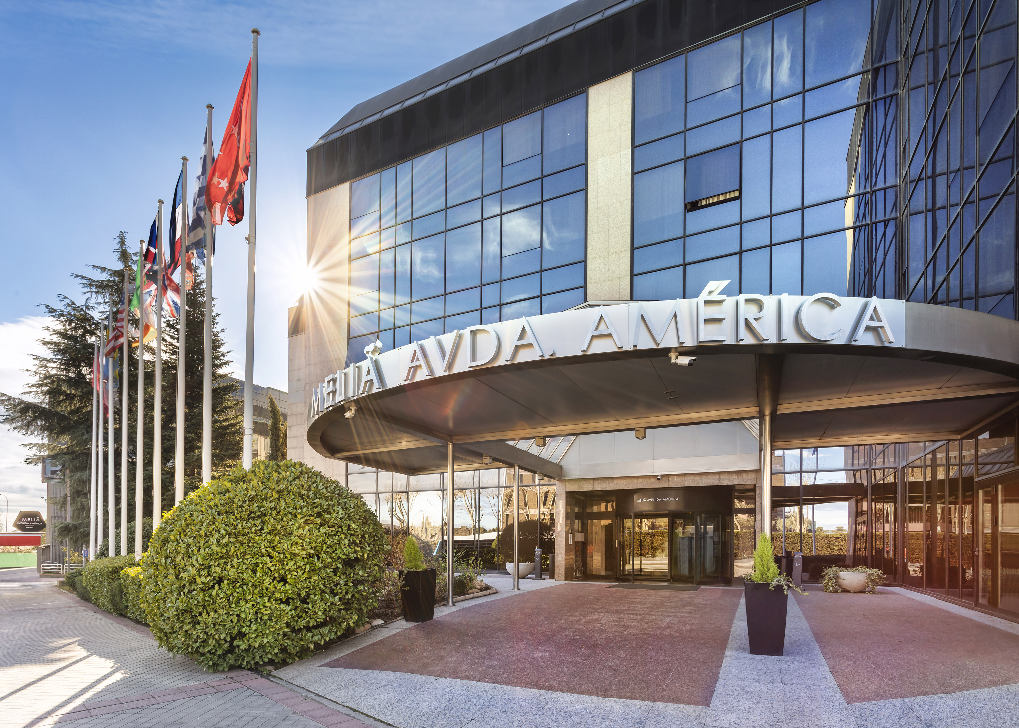 Melia Avda. America hotel entrance, modern glass building, flags, bright sun.