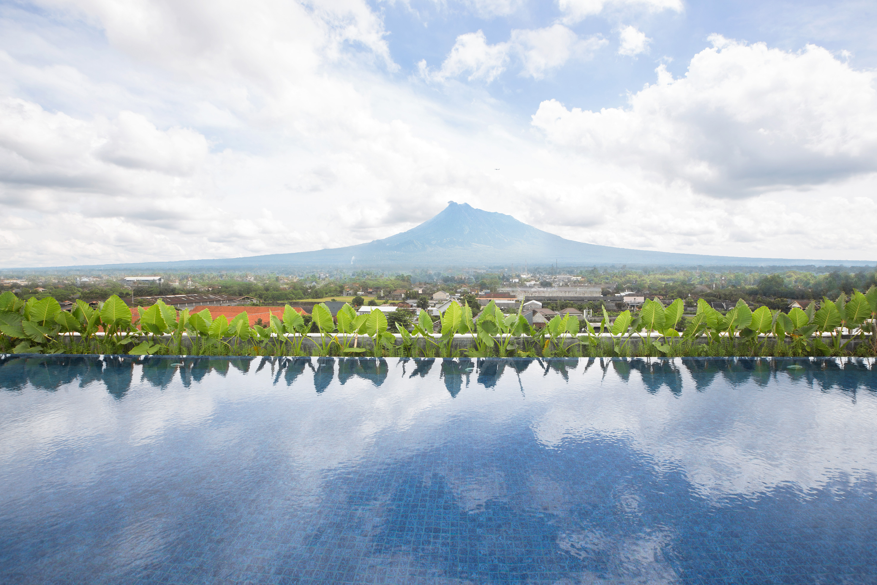 Infinity pool overlooking city, green plants, and distant volcano mountain.