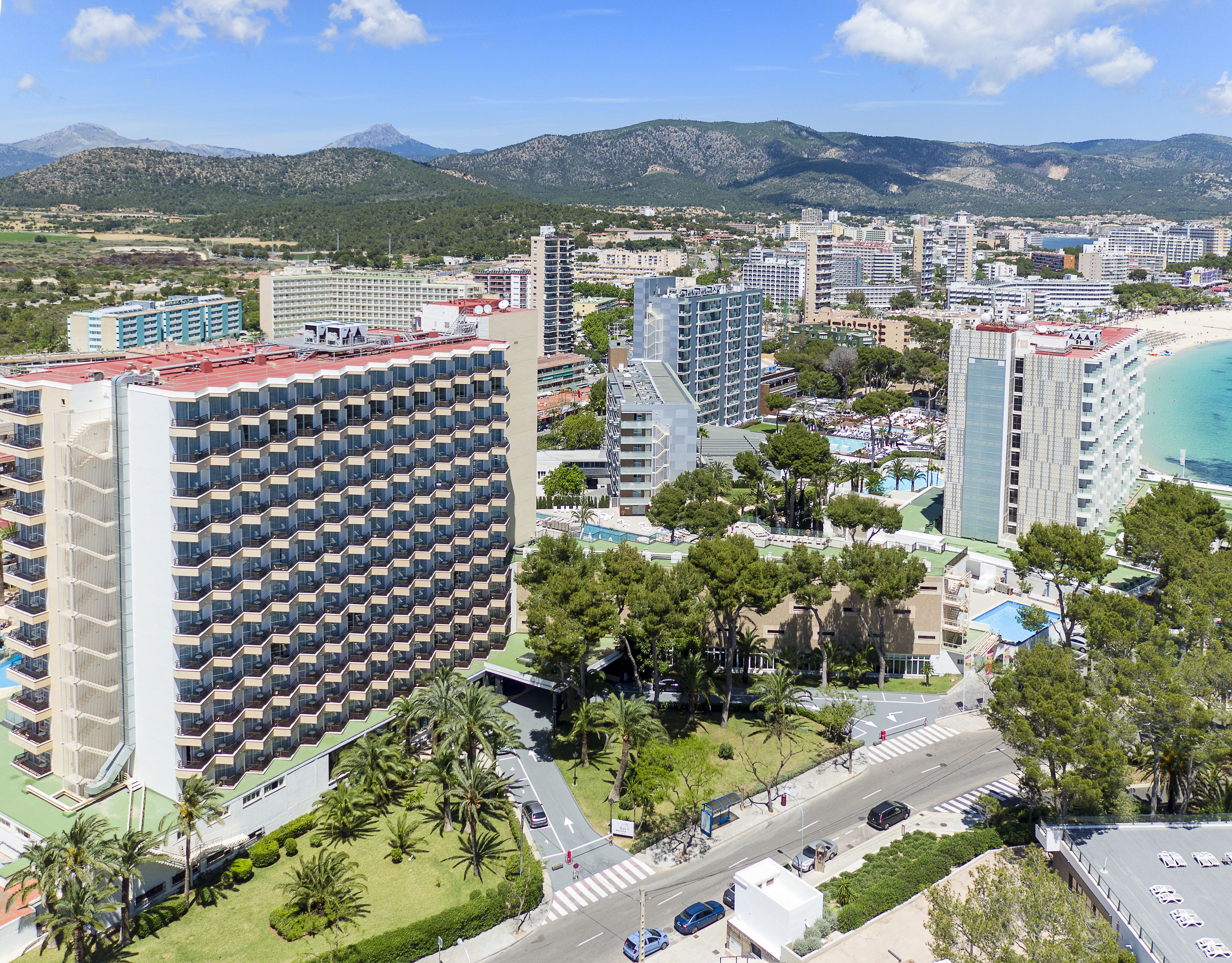 Aerial view: coastal resort, beachfront hotels, turquoise sea, and mountains.