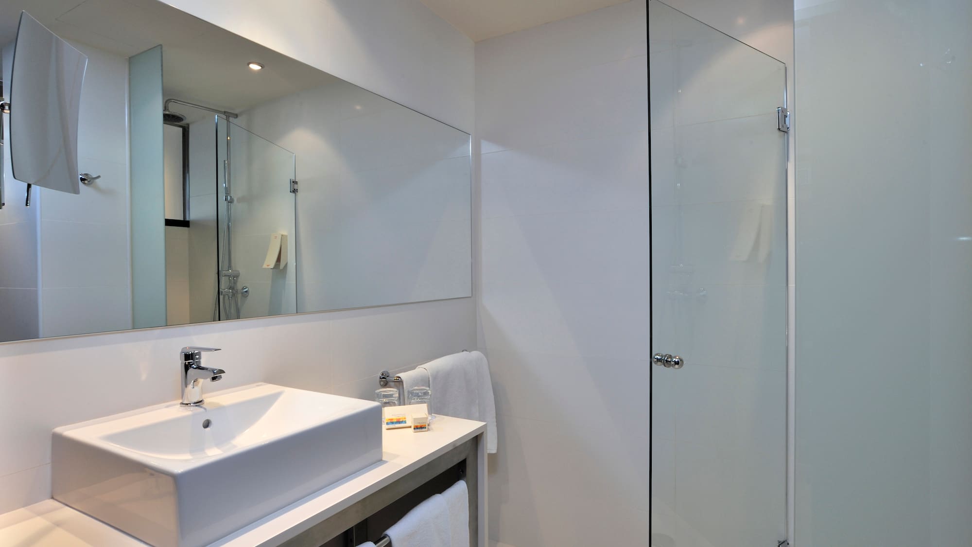Modern white hotel bathroom with vessel sink, large mirror, and glass shower.