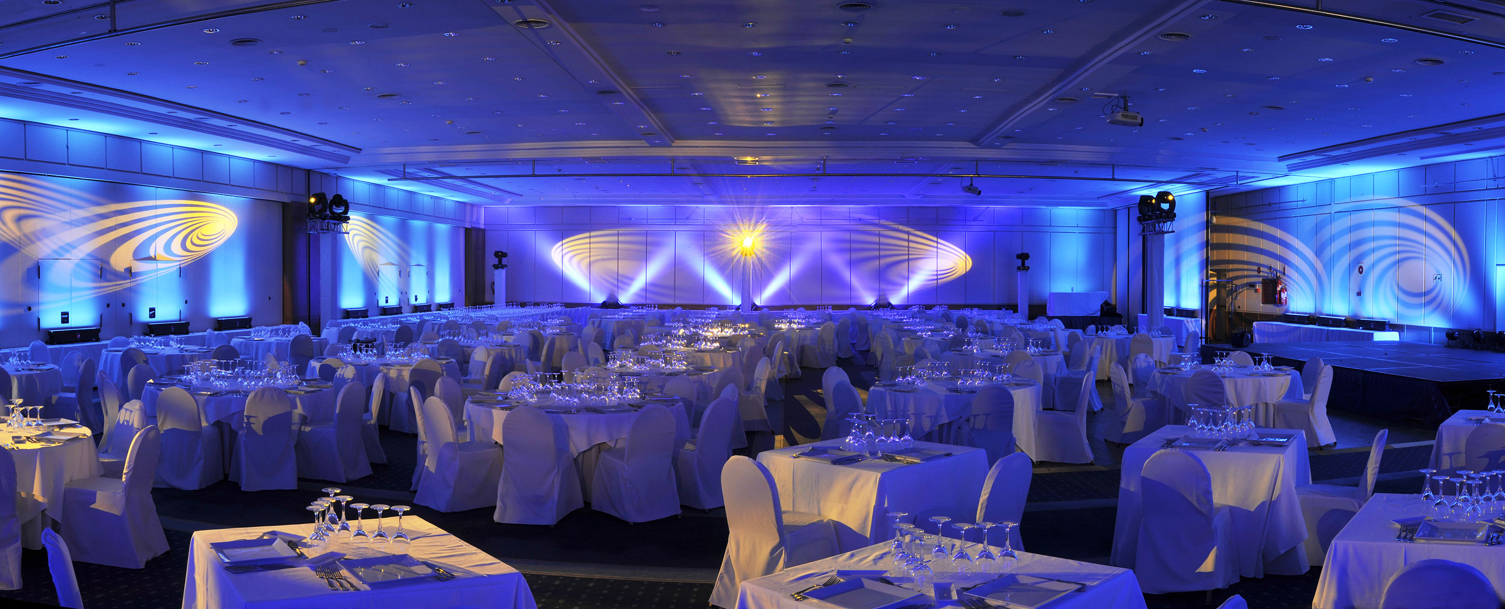 Blue lit ballroom with white tables, chairs, glasses, and dynamic wall projections.