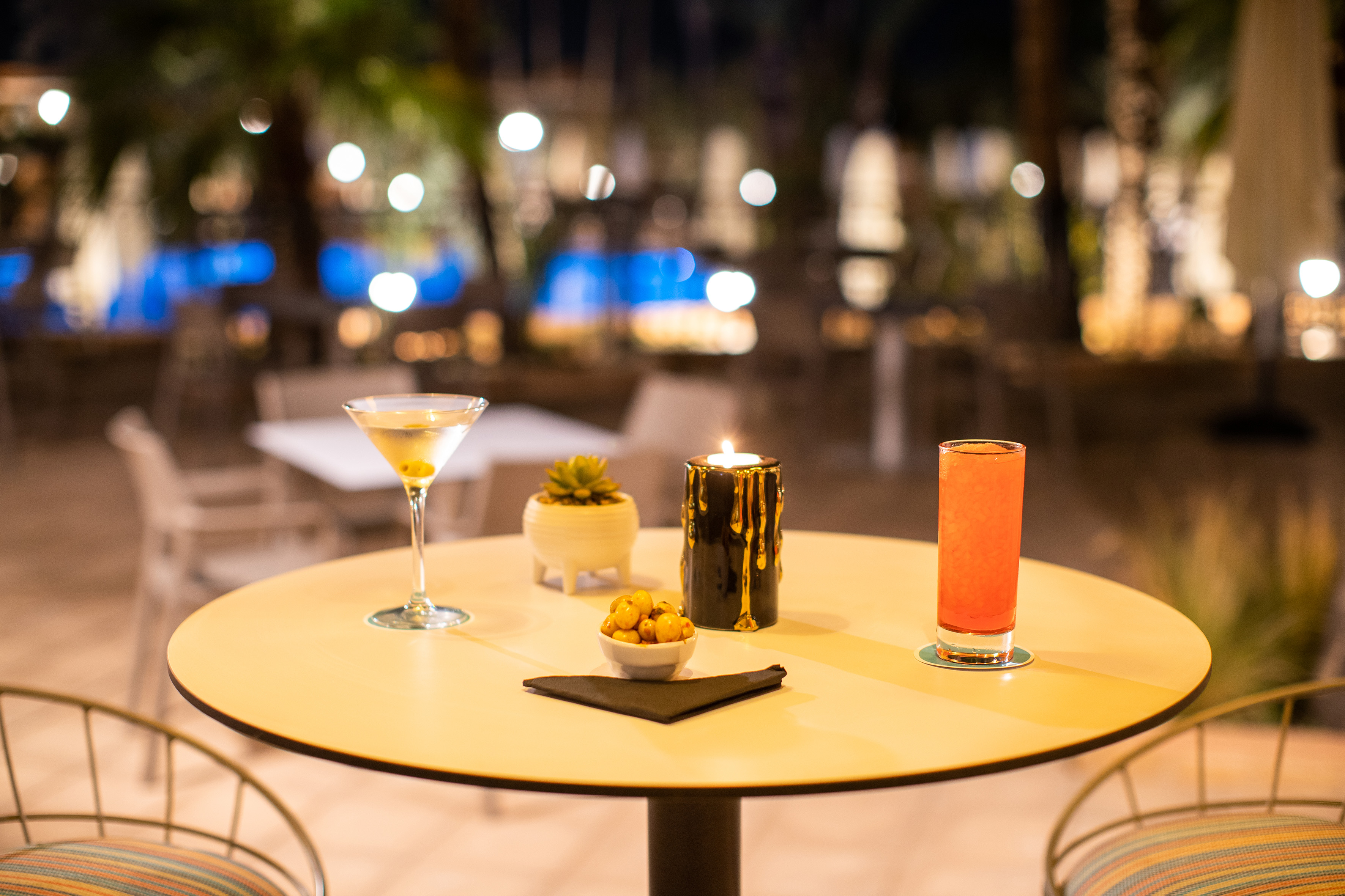 Night bar table with martini, orange cocktail, lit candle, and snacks.