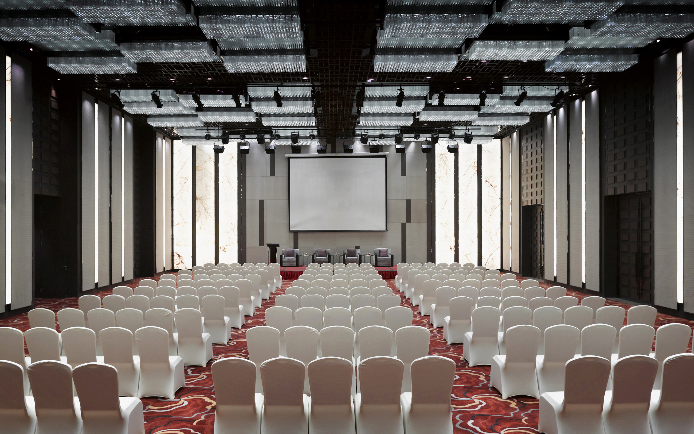 Empty conference room with rows of white chairs, stage, and projector screen.
