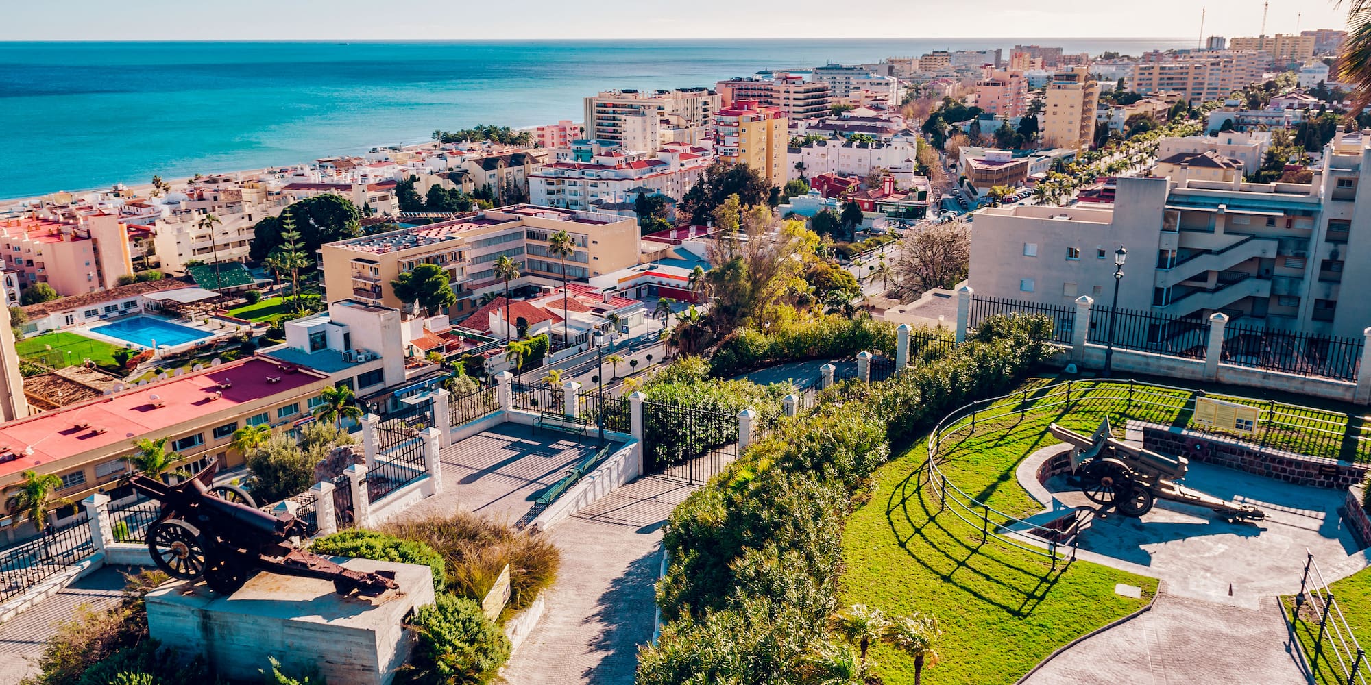 Panorámica costera con mar azul, edificios, playa y cañones en parque.