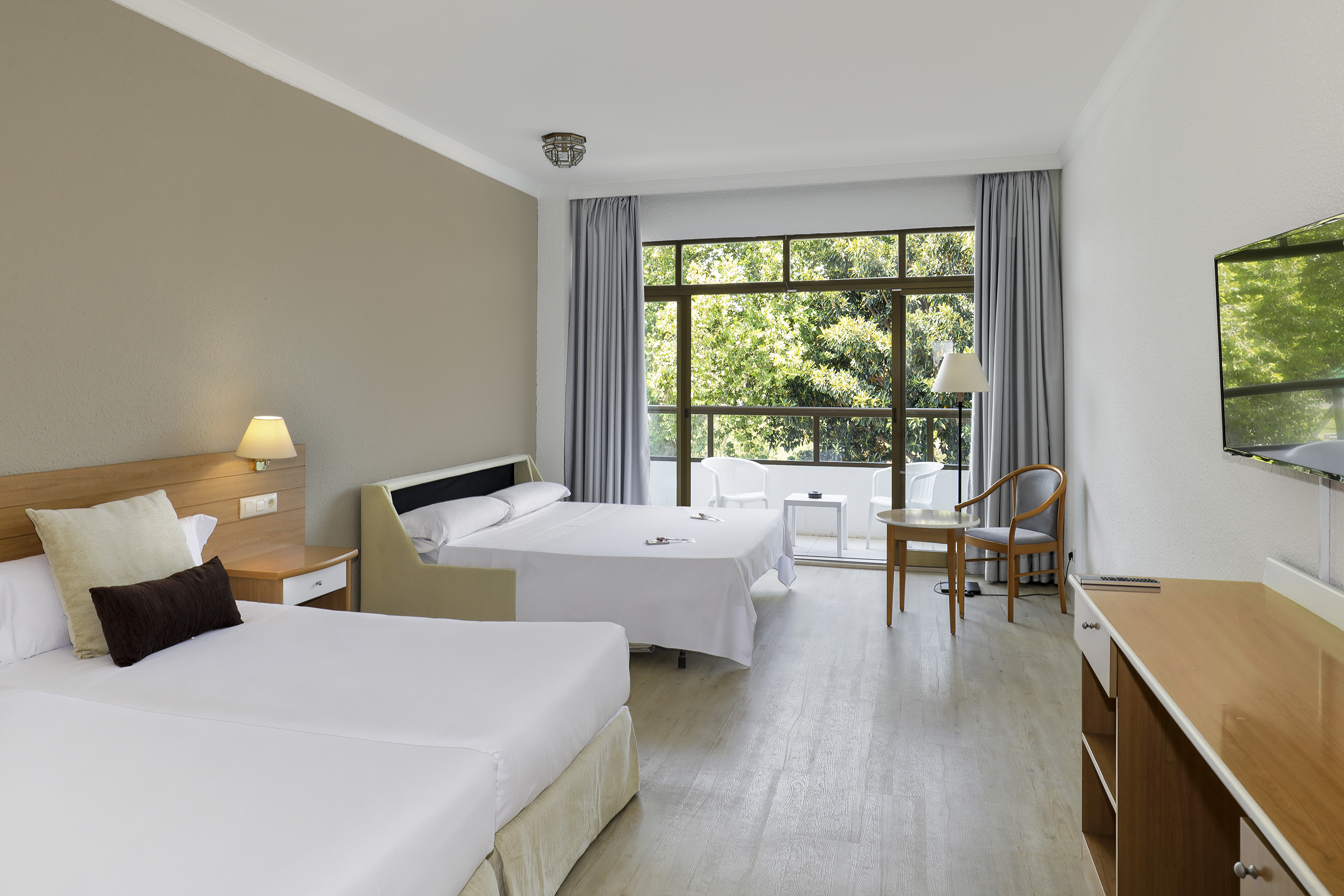 Hotel room with twin beds, sofa bed, balcony and lush green garden view.