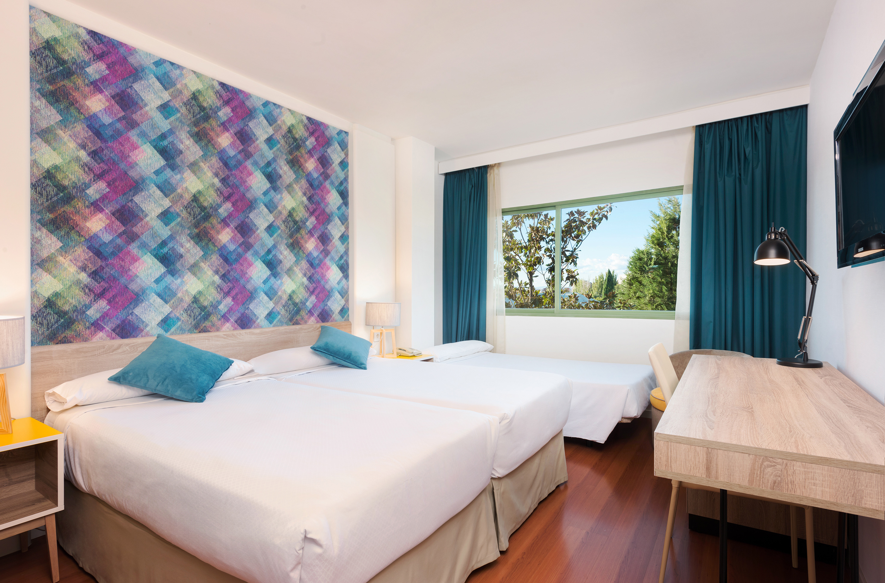 Modern triple hotel room with colorful wall, three beds, desk, and nature view.