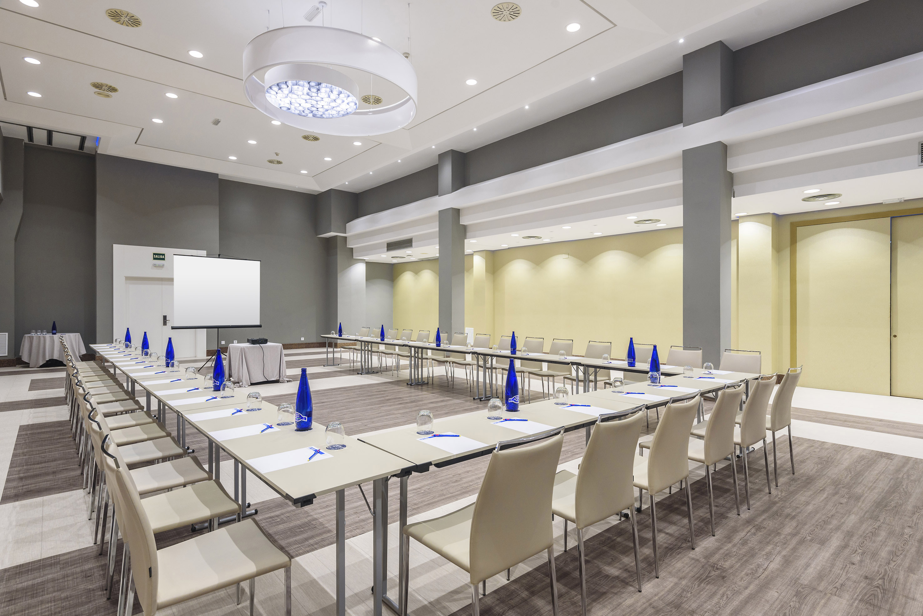 Modern conference room with U-shaped tables, projector screen, and beige chairs.
