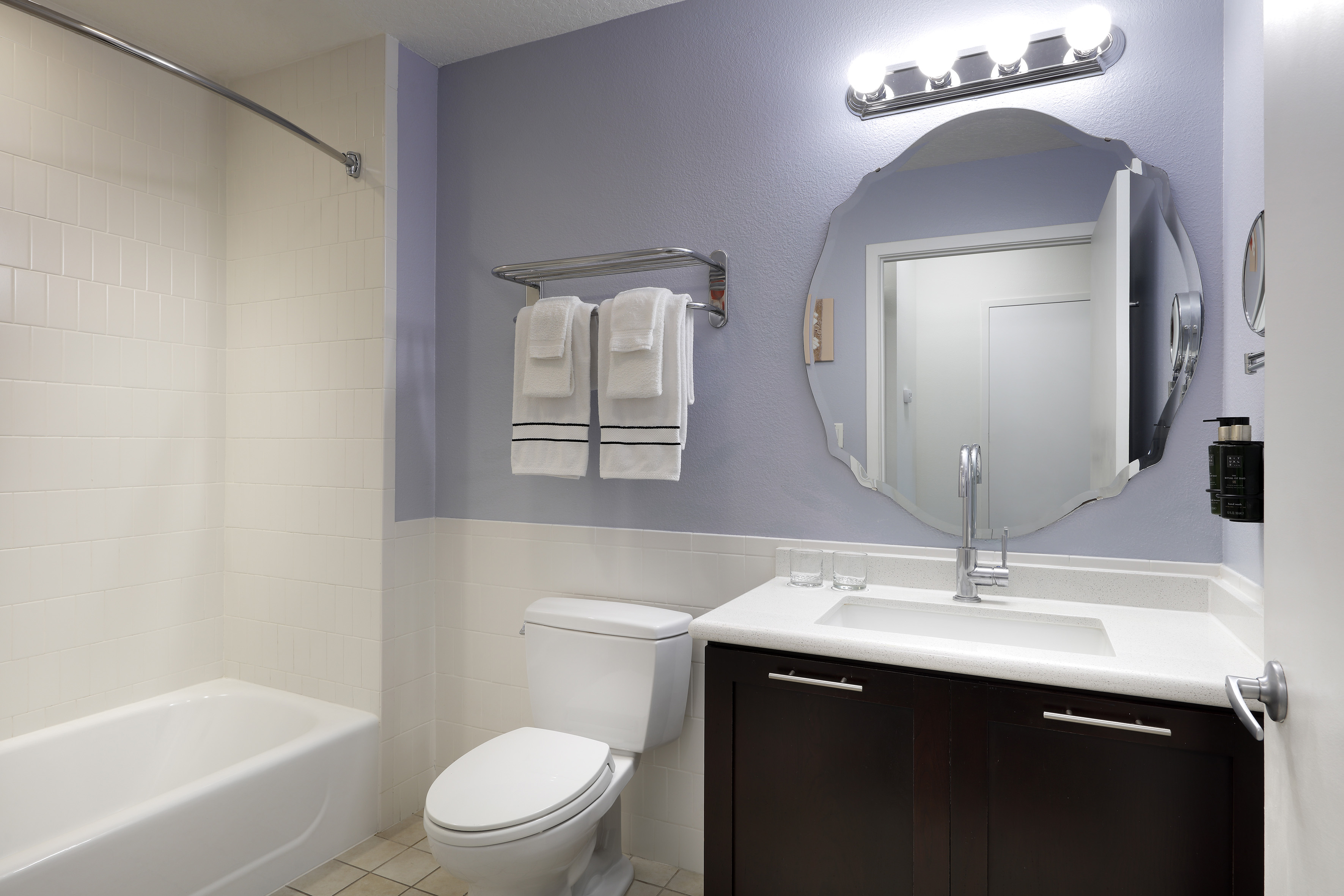 Clean hotel bathroom with bathtub, toilet, dark vanity, mirror, and lilac walls.