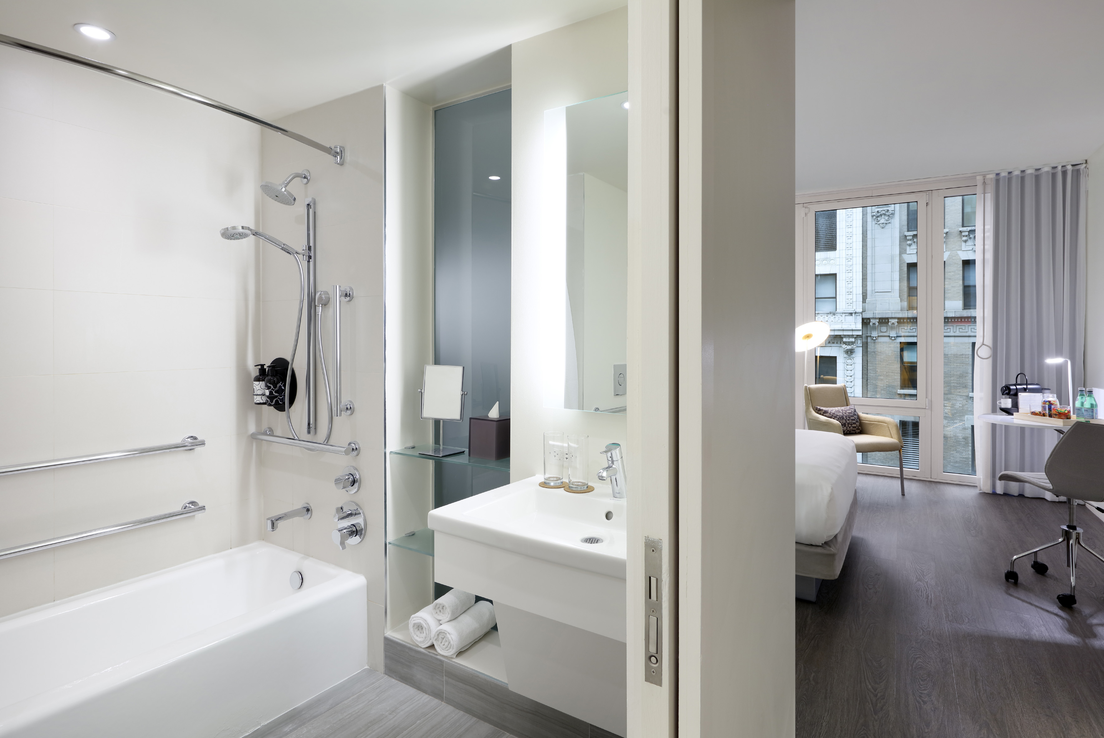 Modern hotel bathroom with white tub, shower, sink, and glimpse of bedroom.