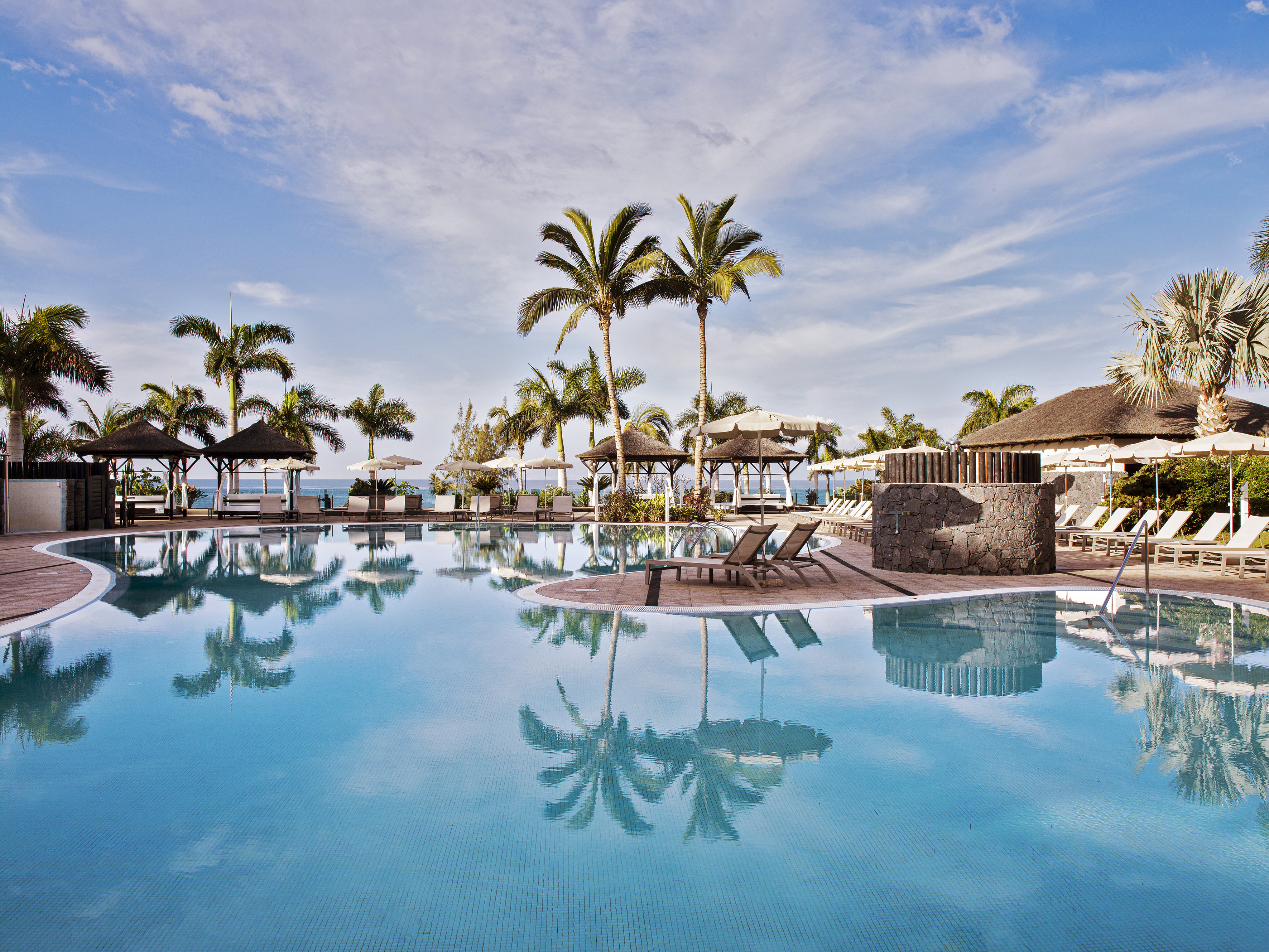 Tropical resort swimming pool with palm trees, sun loungers, and cabanas.