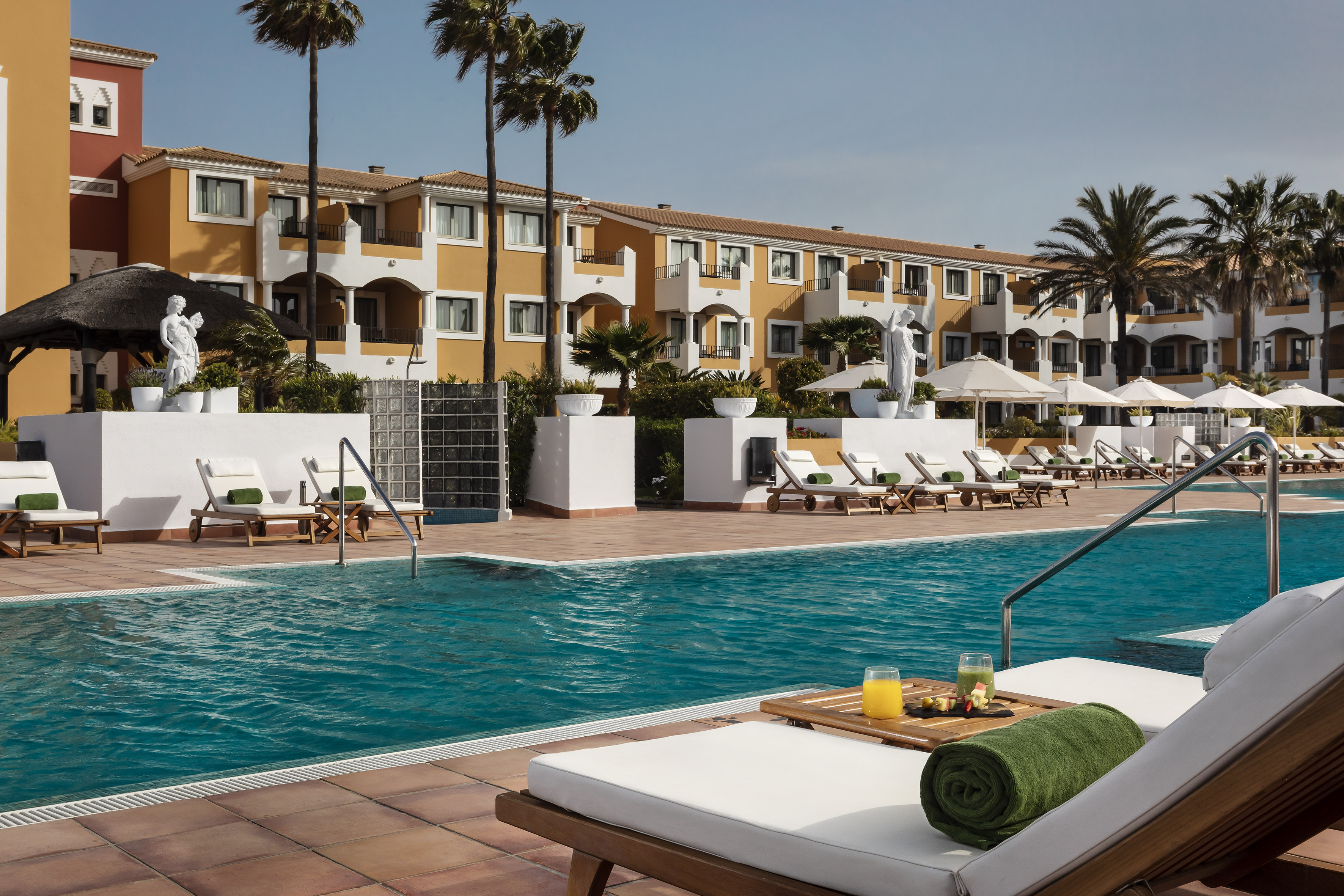 Sunny luxury resort pool, orange hotel, palm trees, and relaxing loungers.