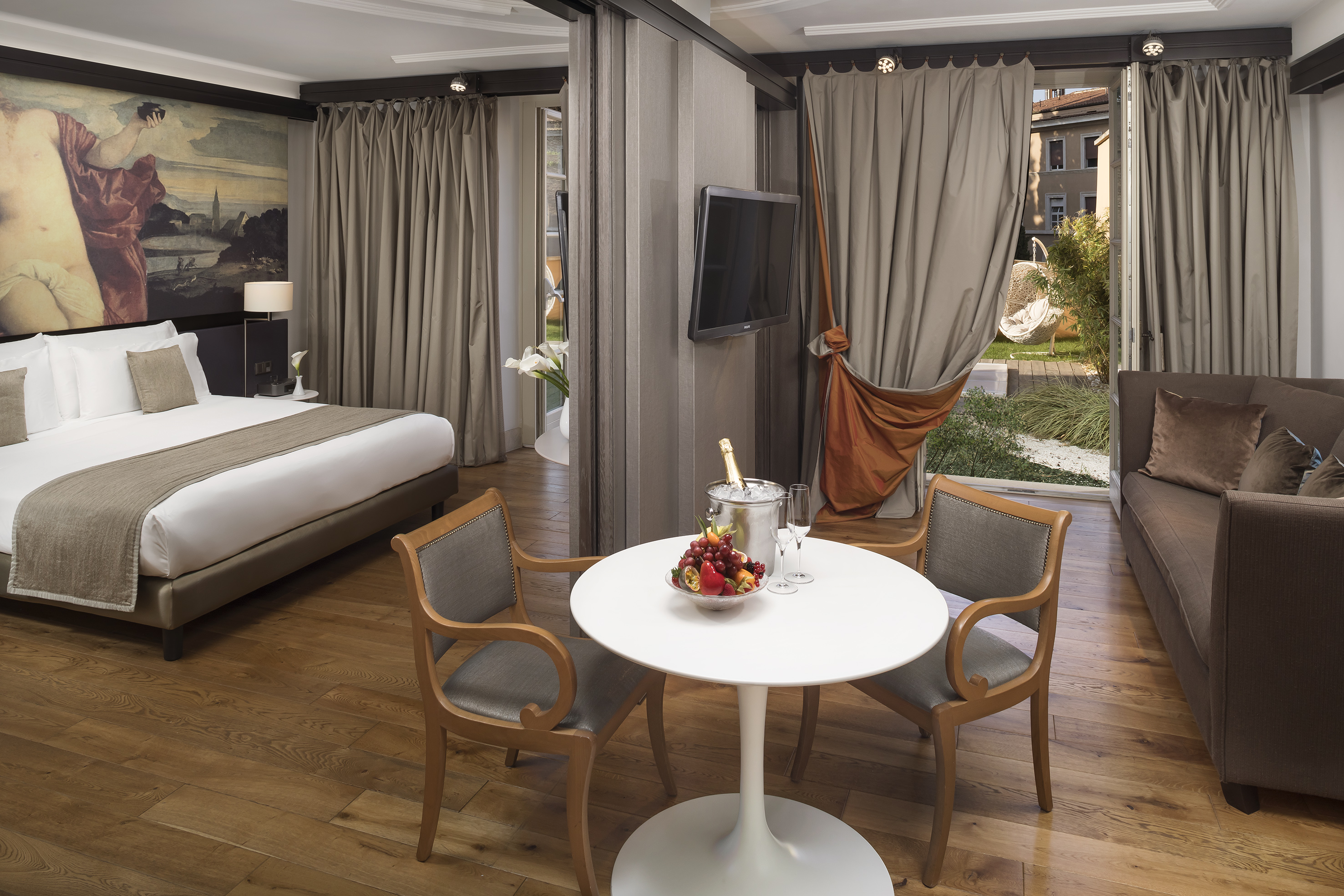 Elegant hotel suite: bed, sofa, dining, champagne, fruit, garden view.