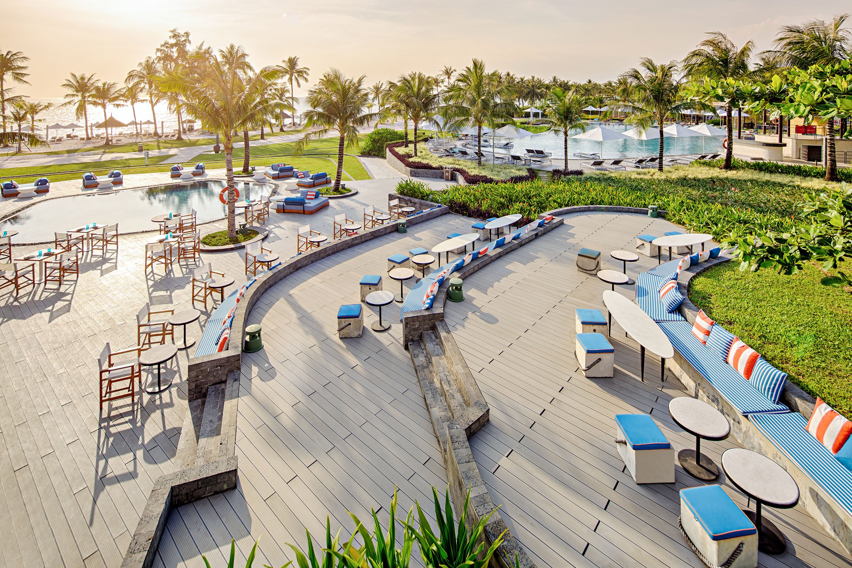 Tropical resort poolside lounge, outdoor dining, palm trees, sunset.