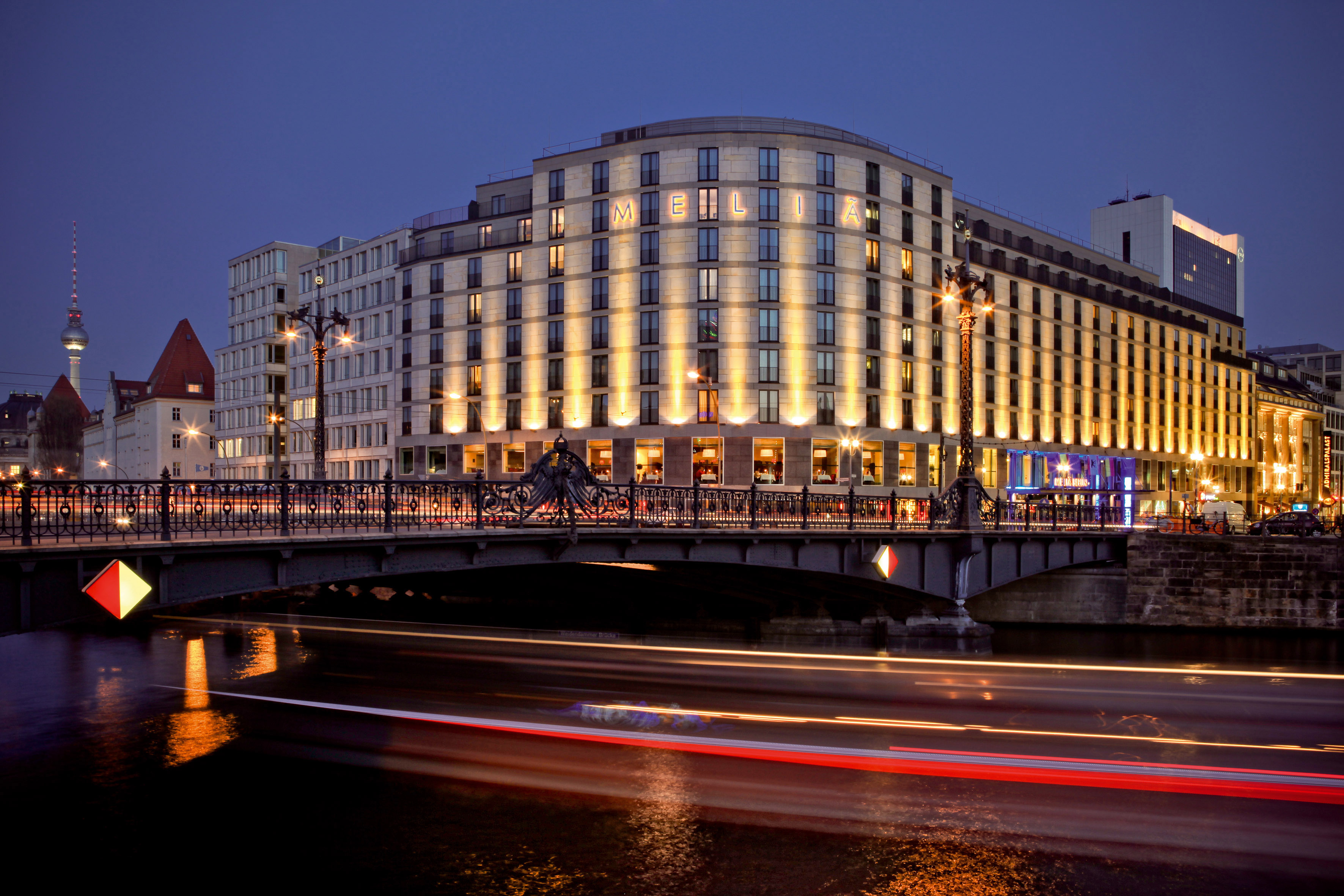 Illuminated Meliá Berlin hotel and bridge over river at night, city light trails.