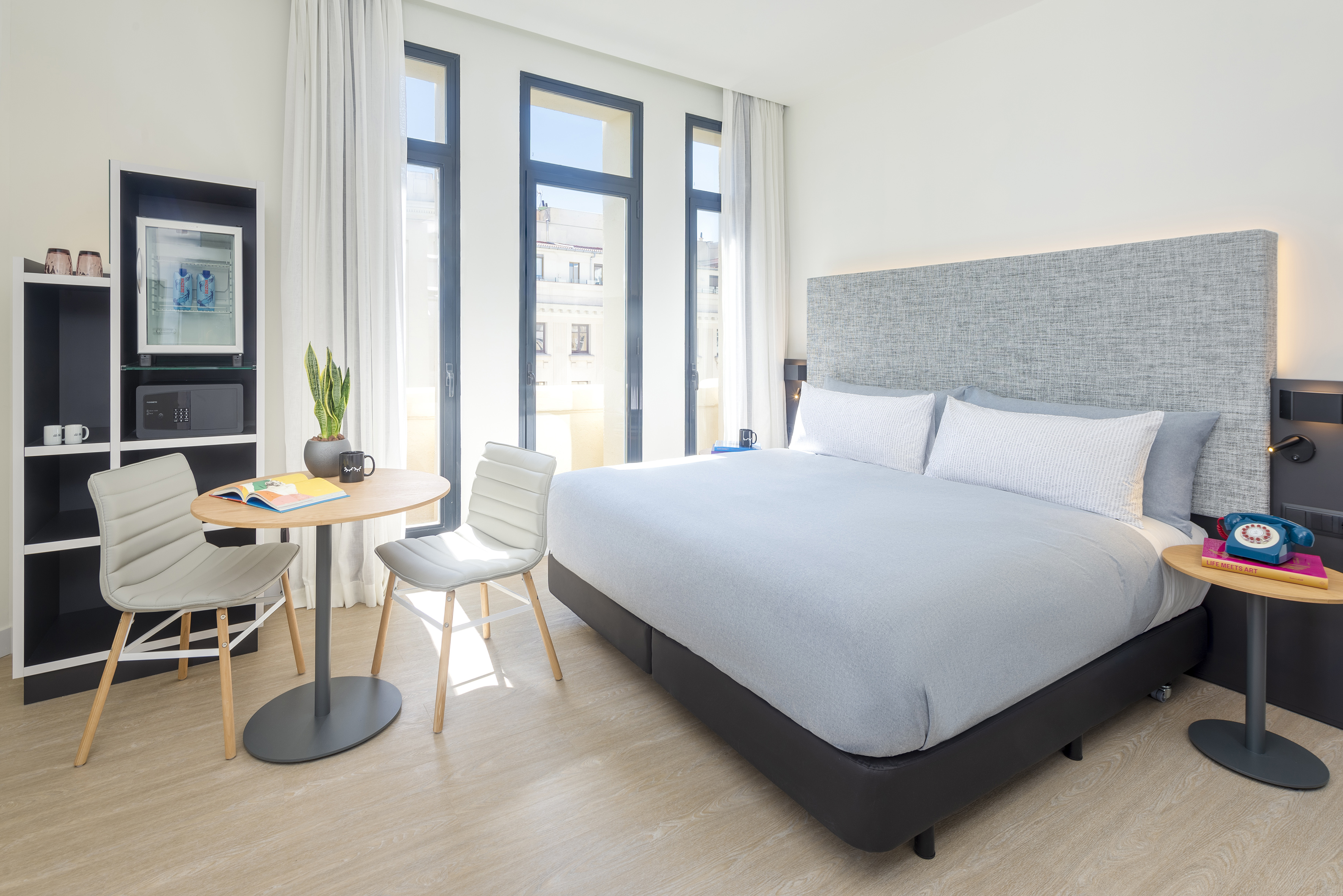 Modern hotel room with large bed, floor-to-ceiling windows, and dining table.