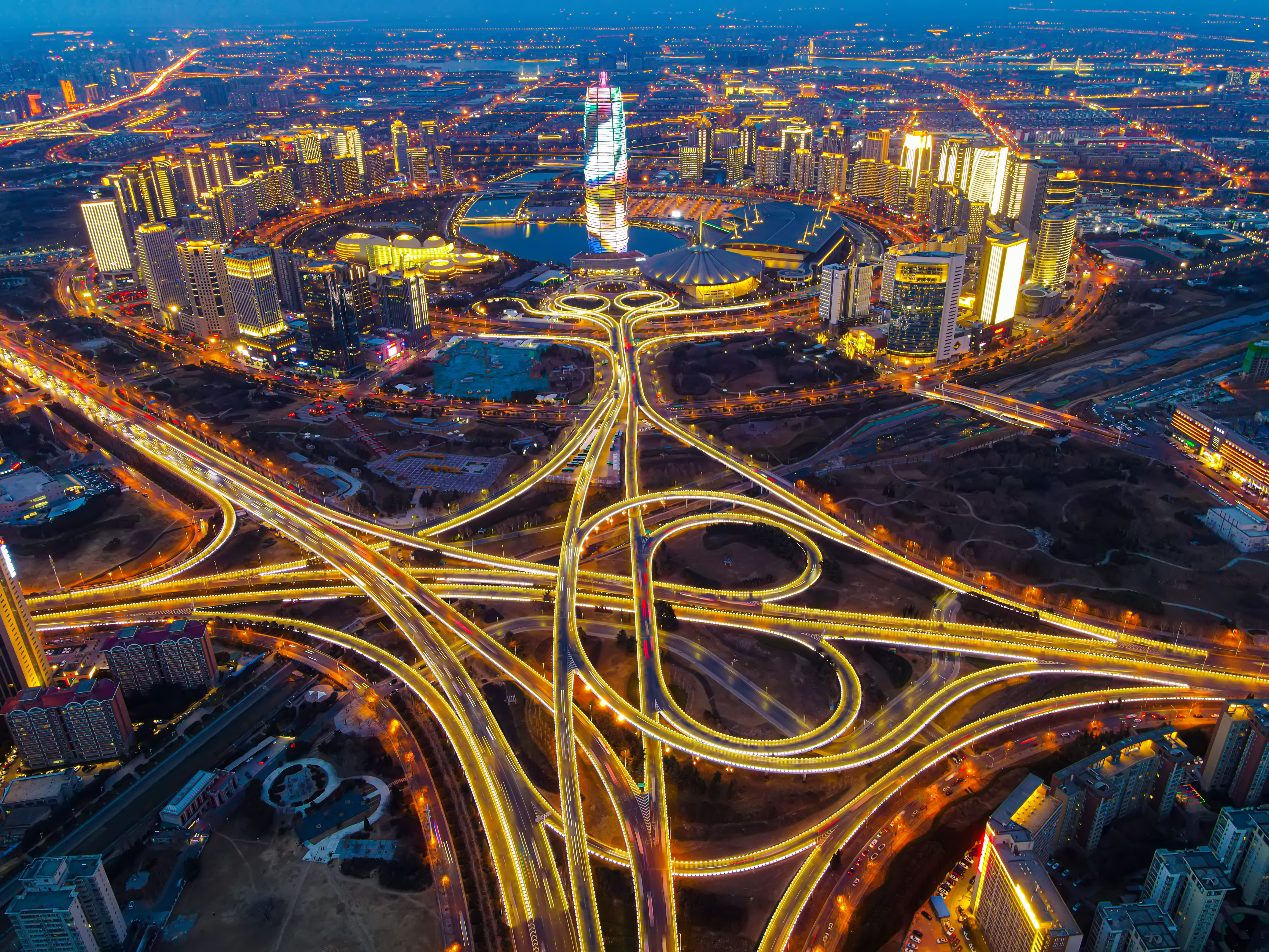 Night city aerial: spiraling tower, bright skyline, complex highway lights.