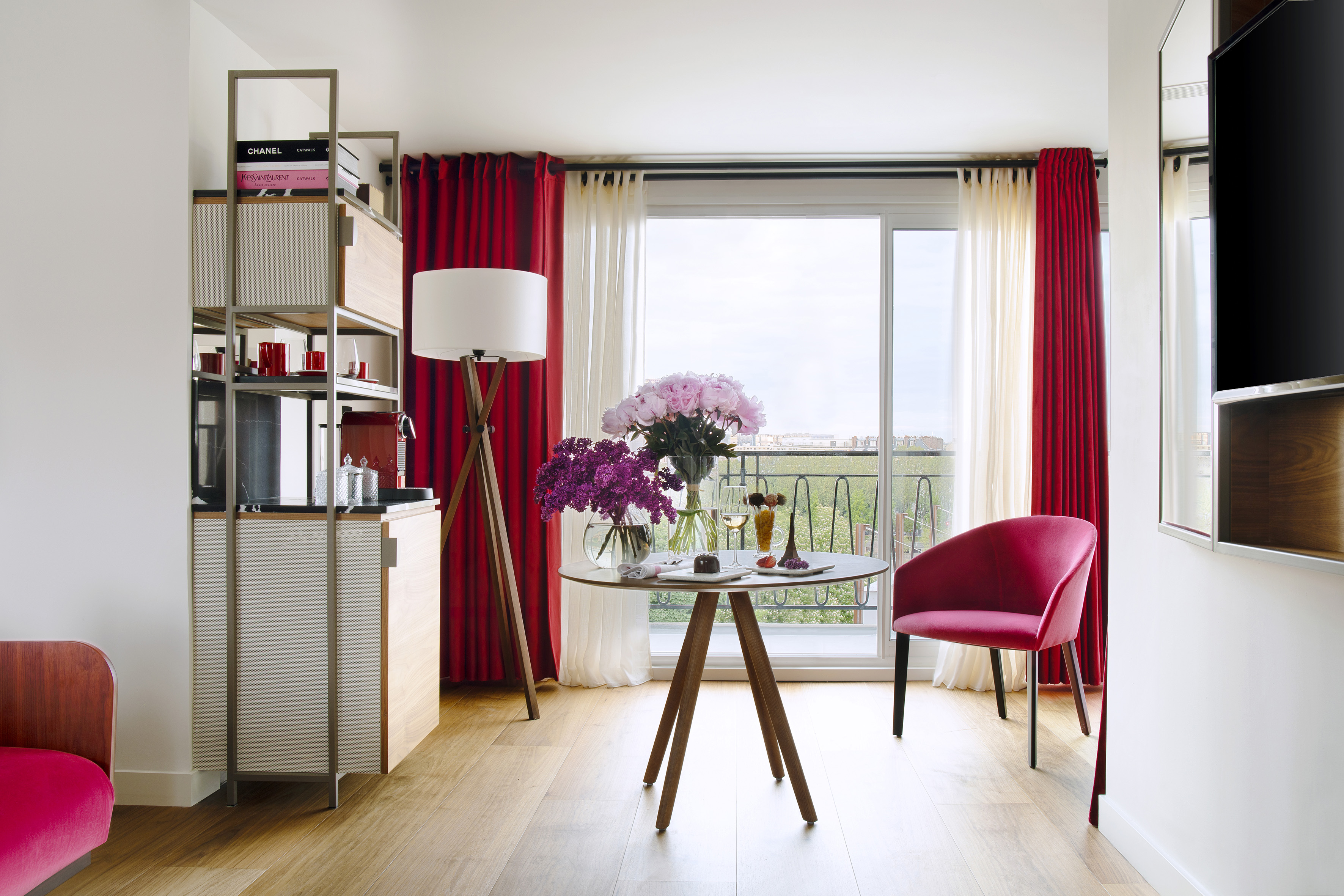 Chic hotel suite with balcony, red drapes, pink seating, and elegant decor.
