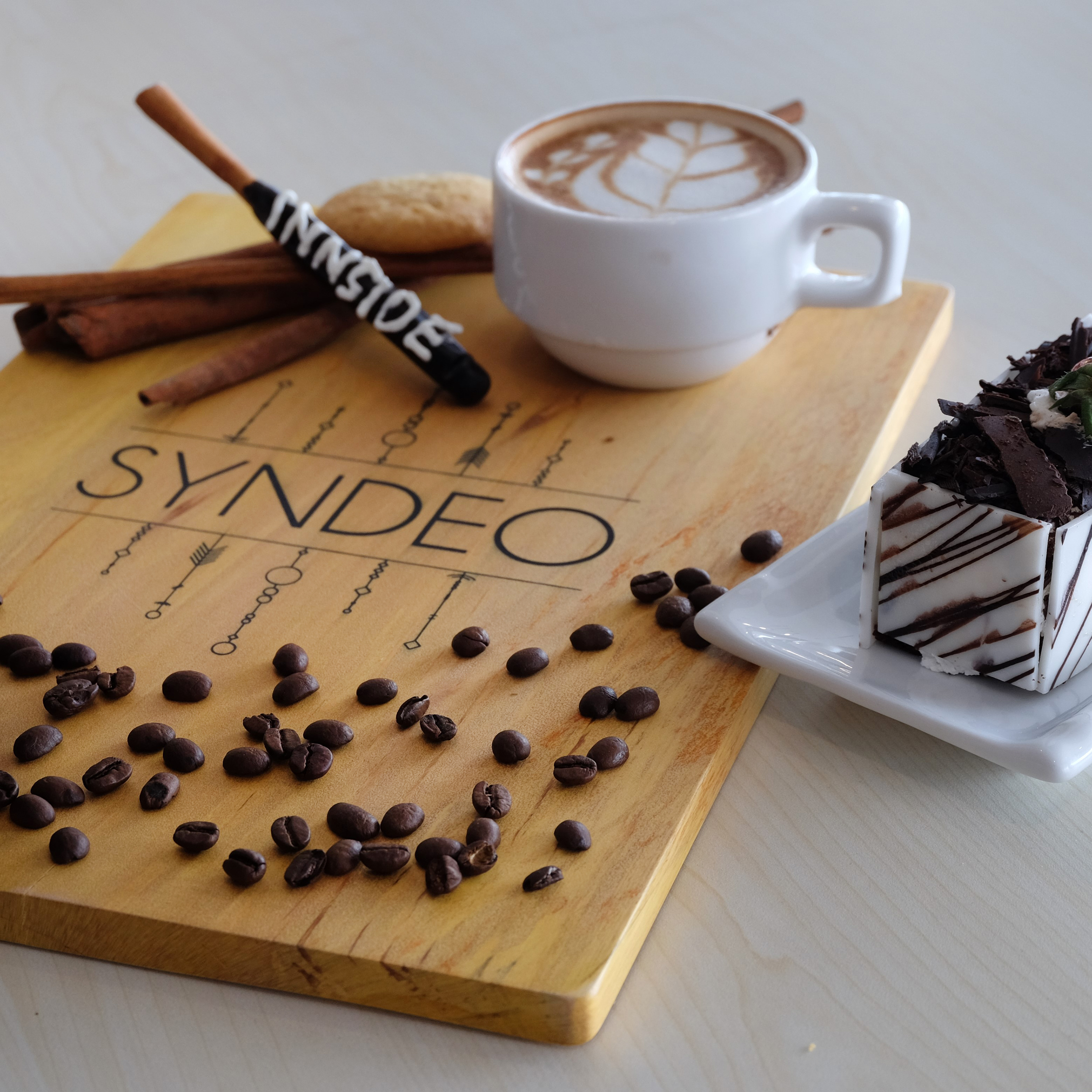 Latte with art, chocolate cake, and coffee beans on a SYNDEO wooden board.