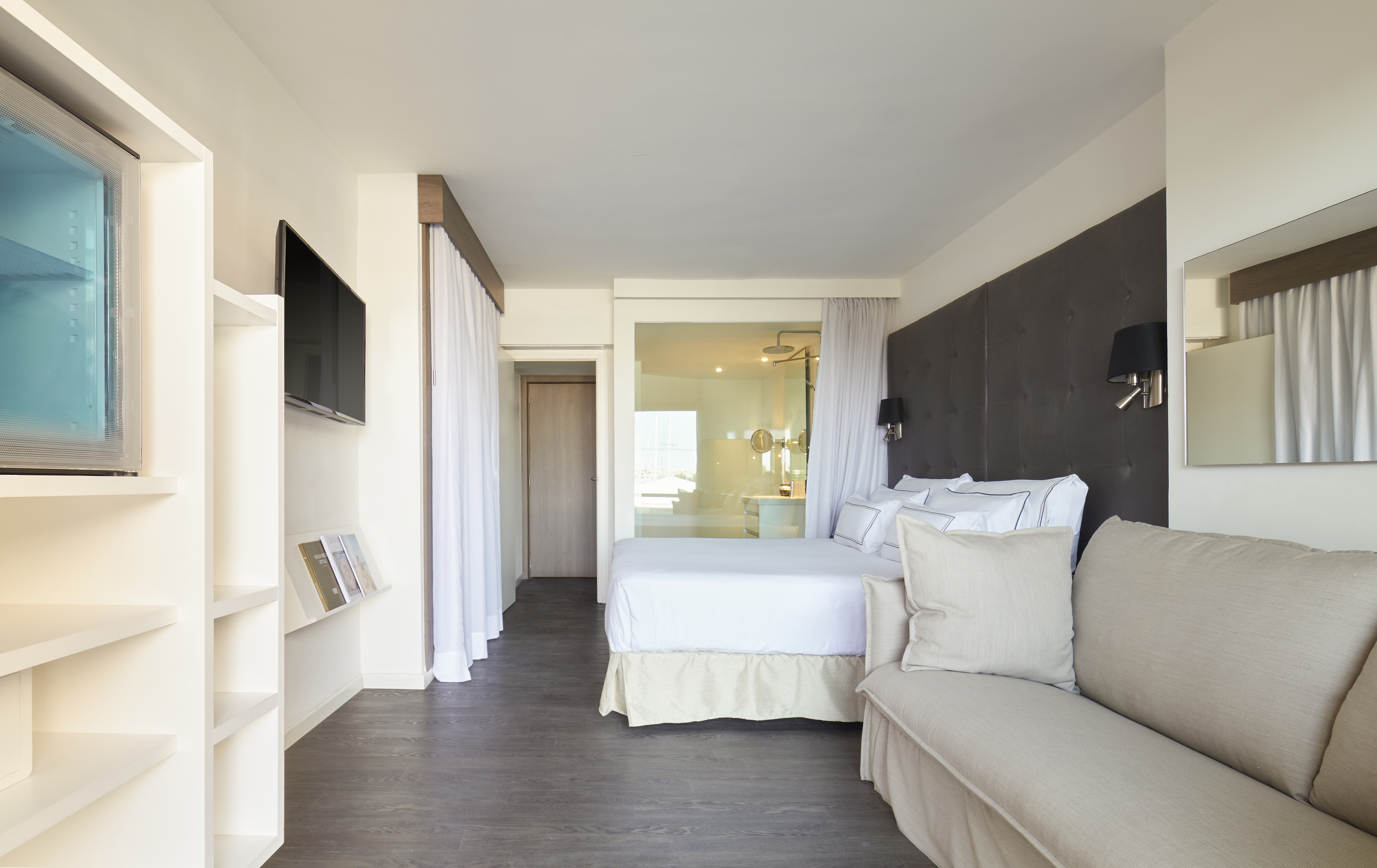 Modern hotel room with king bed, sofa, TV, and open glass bathroom.