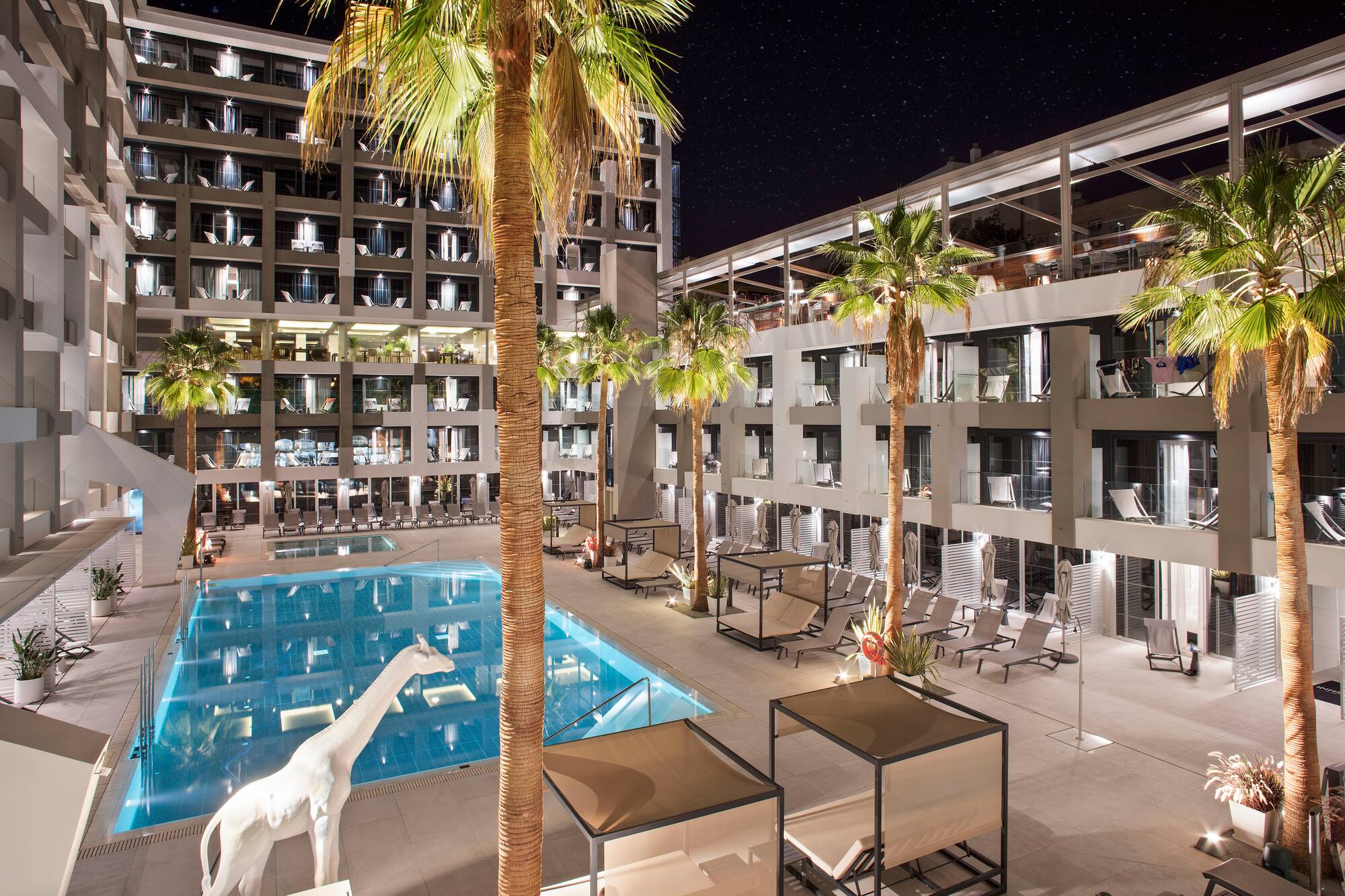 Night hotel resort pool, palm trees, illuminated balconies, white giraffe statue.