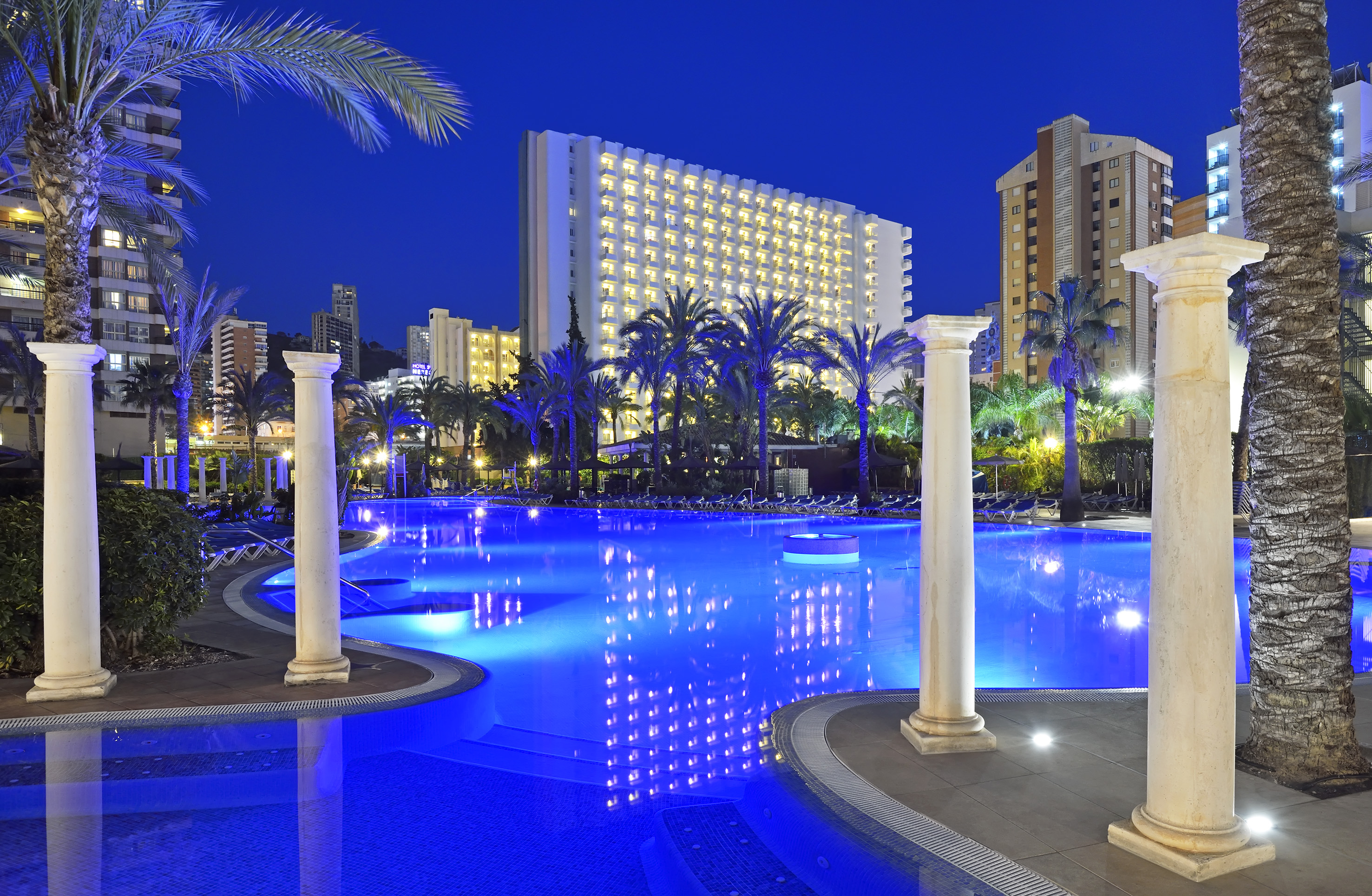Illuminated hotel pool at night with palm trees and tall buildings.