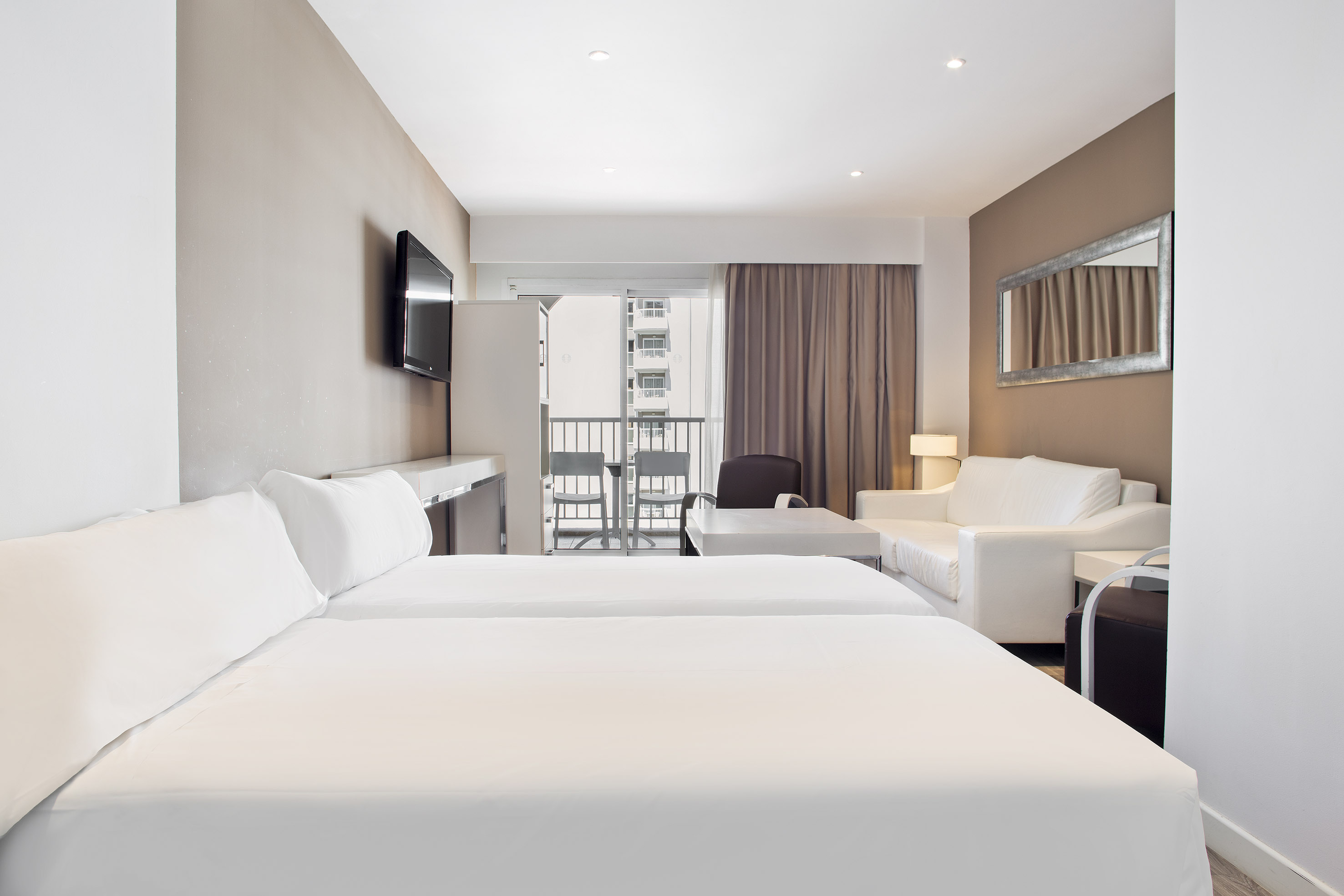 Modern hotel studio with twin beds, sofa, wall TV, and city balcony.