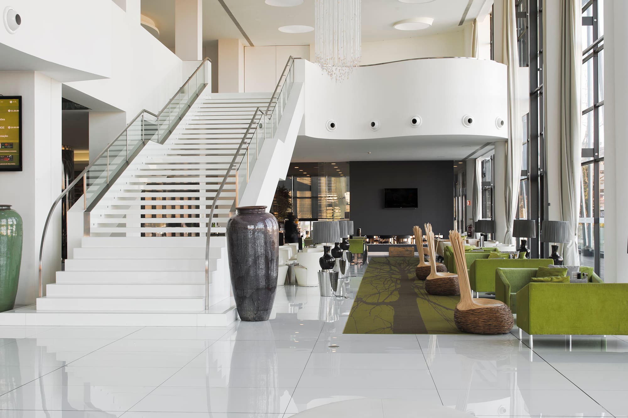 Bright hotel lobby with white staircase, crystal chandelier, and green seating.