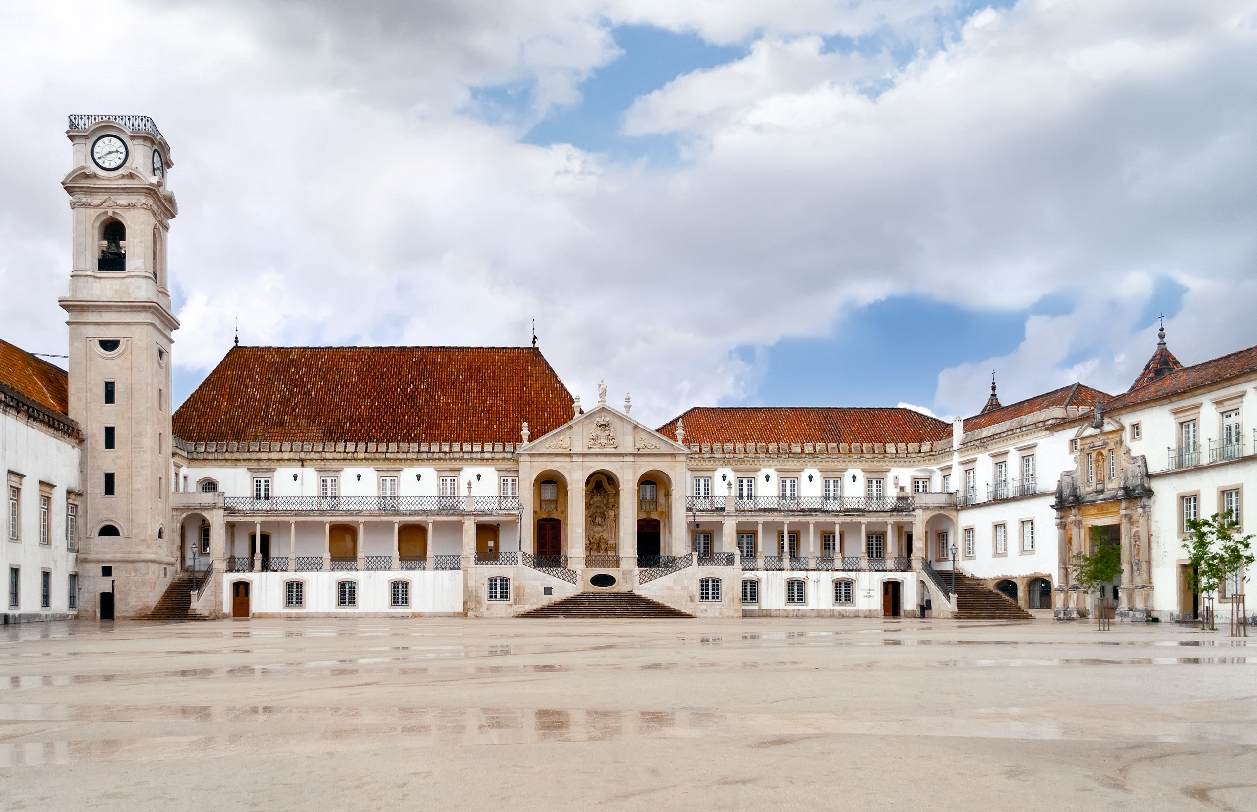 University of Coimbra historic palace, iconic clock tower, and red roofs on wet square.
