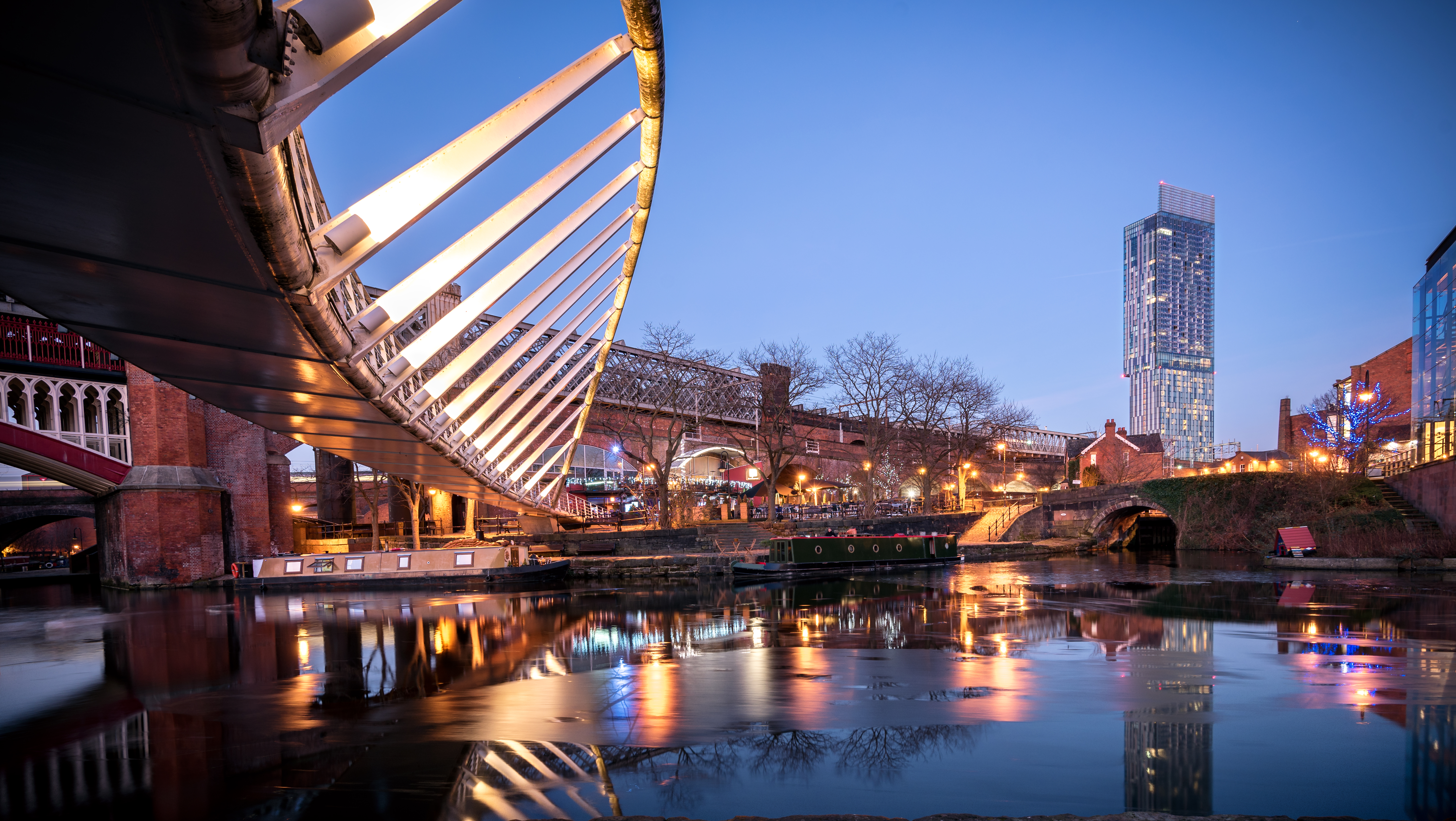 Dusk over Manchester canal with illuminated bridge, boats, and city skyscraper.