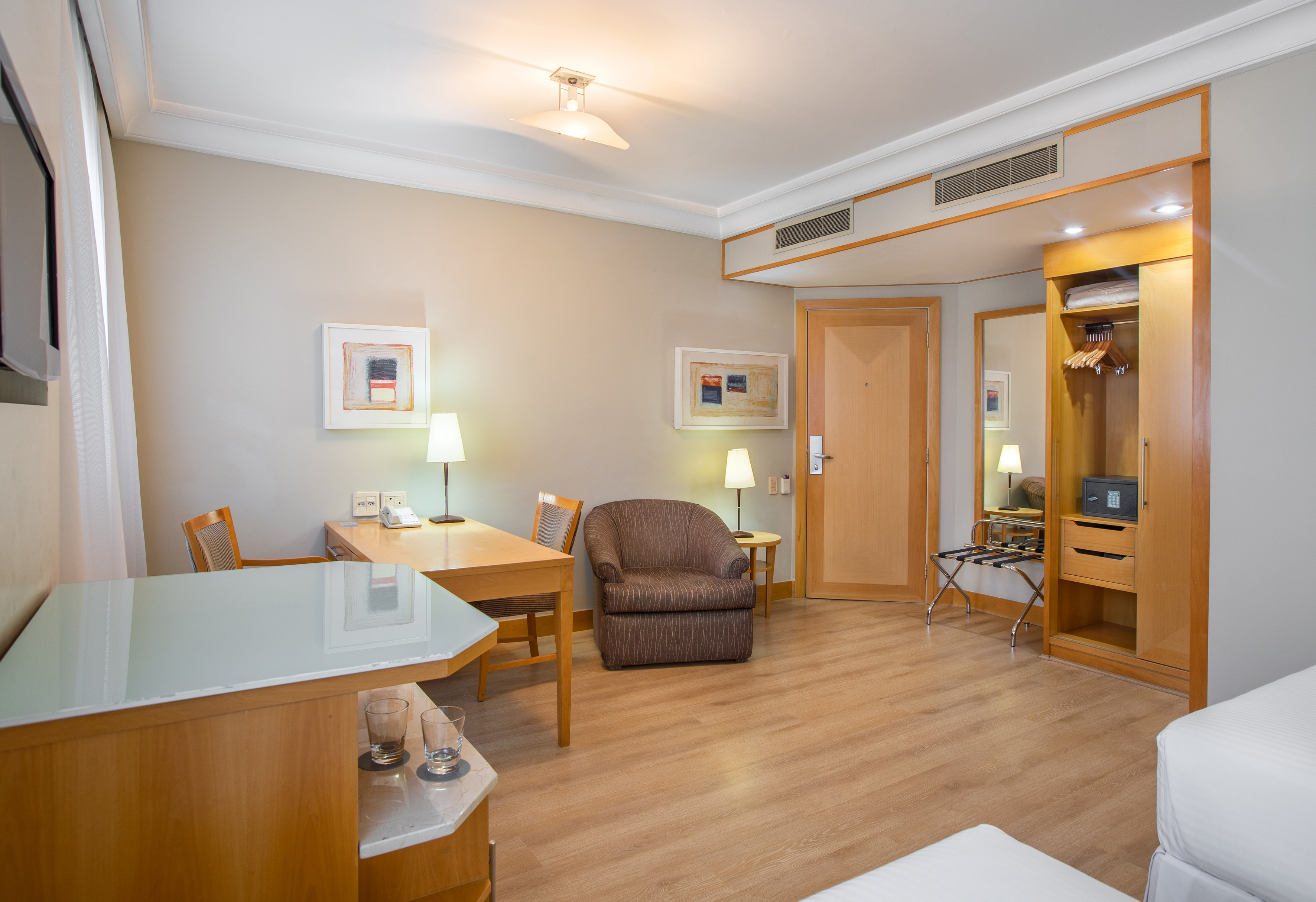 Hotel room with light wood floor, work desk, brown armchair, and open closet.