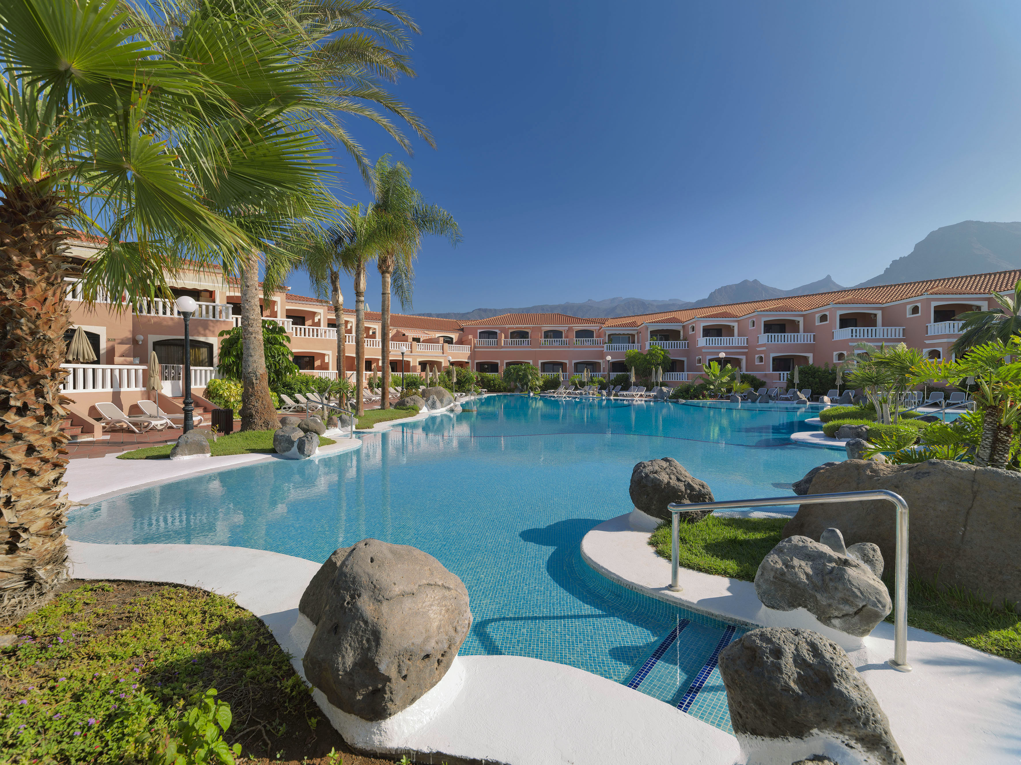 Tropical resort pool with hotel, palm trees, and distant mountains.