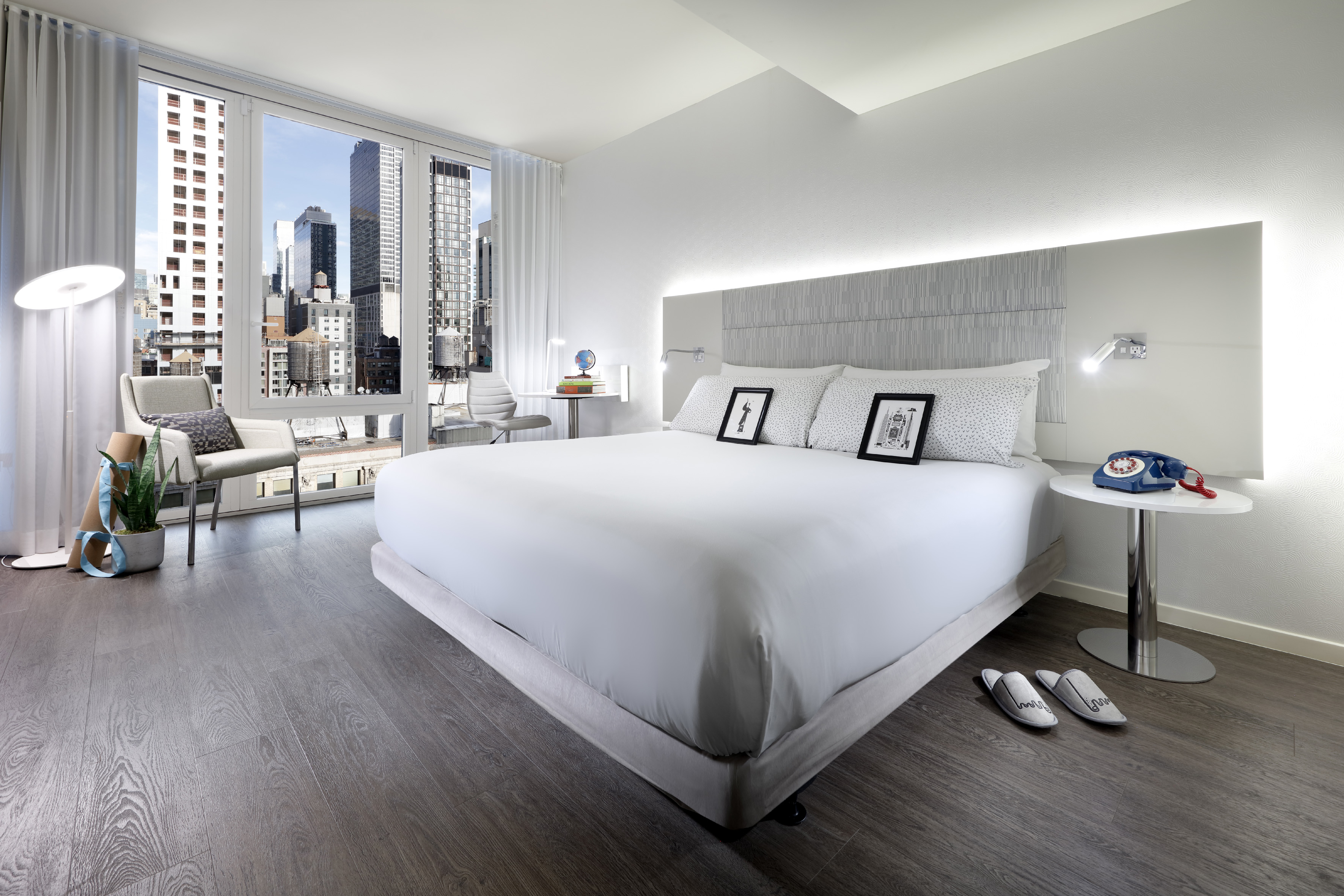 Modern hotel room with city skyline view, white bed, and dark hardwood floor.