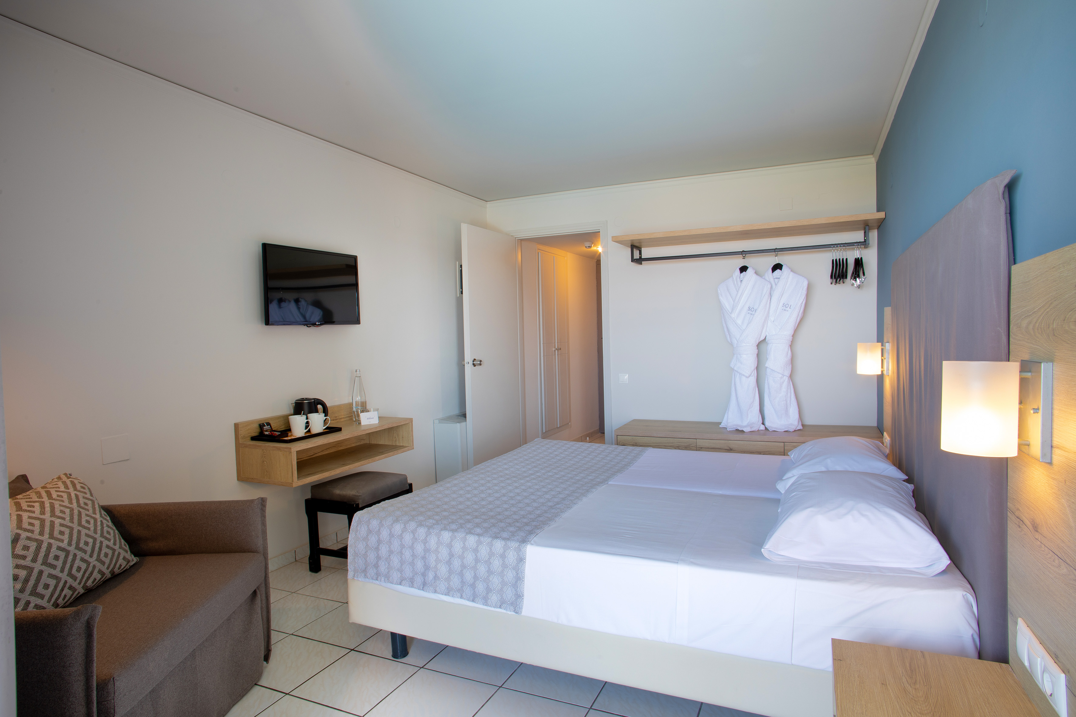 Modern hotel room with twin beds, blue accent wall, wall-mounted TV, and bathrobes.