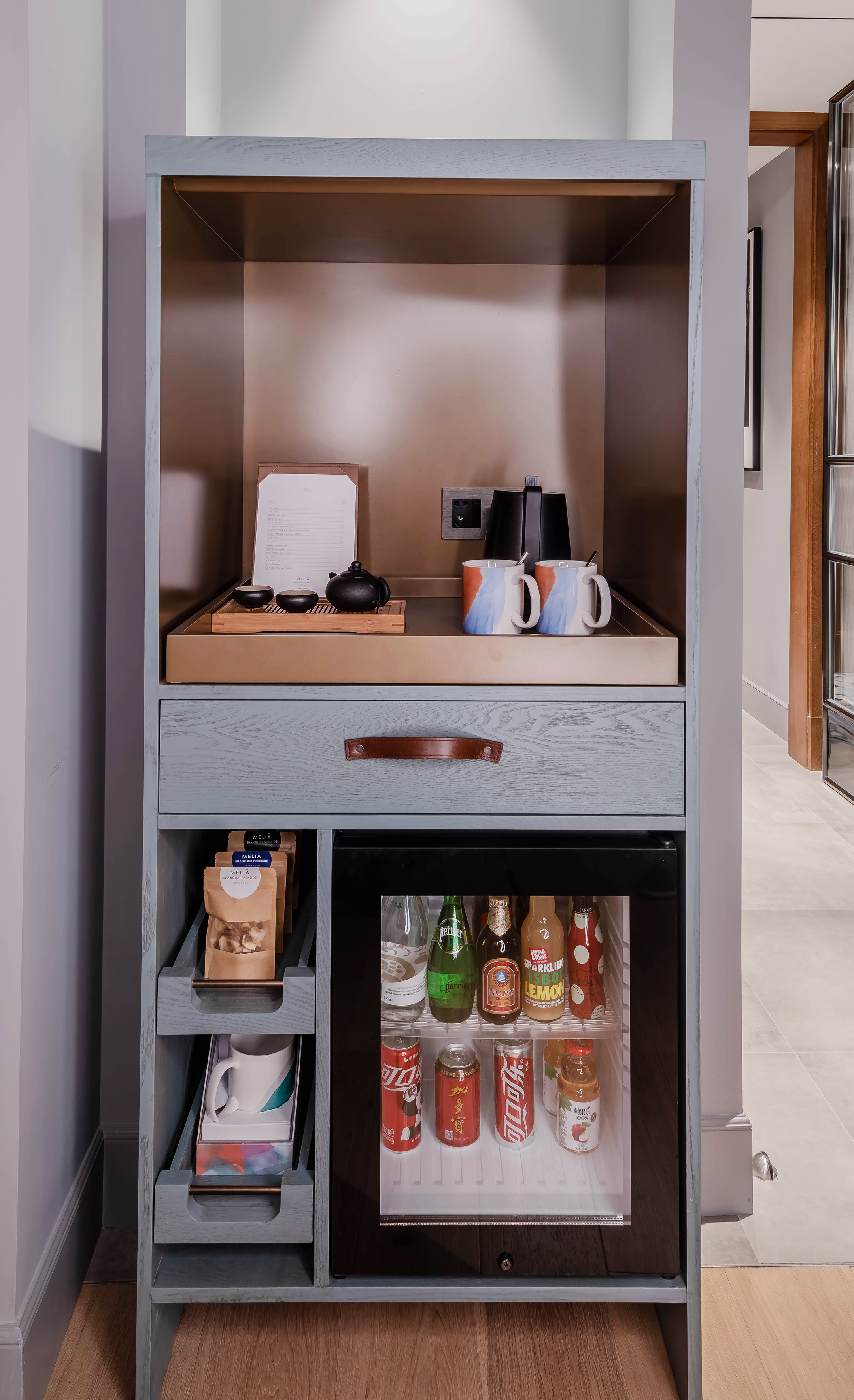 Hotel mini-bar cabinet with tea set, snacks, and assorted beverages.