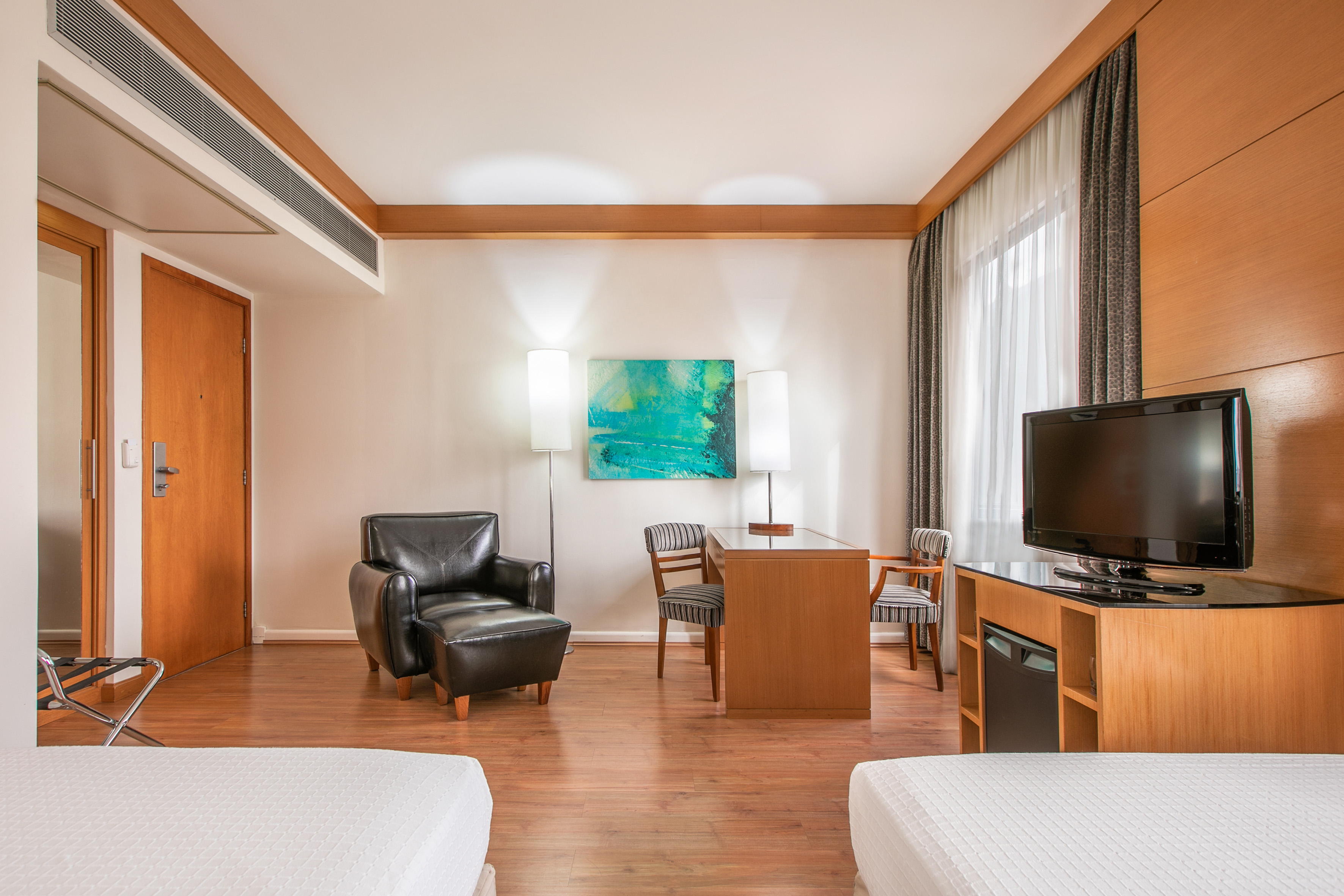 Modern hotel guest room with armchair, work desk, TV, and wood floor.