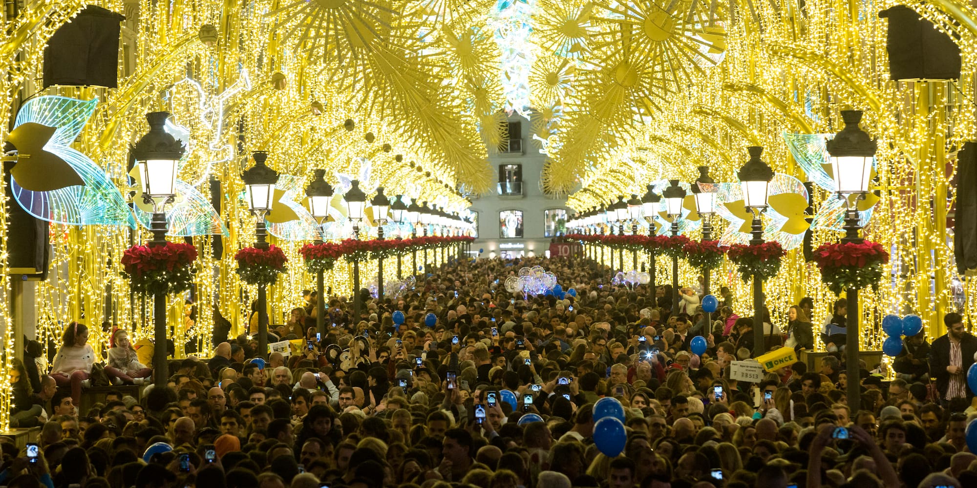 Gran multitud en calle decorada con luces de Navidad doradas. Farolas con poinsettias.