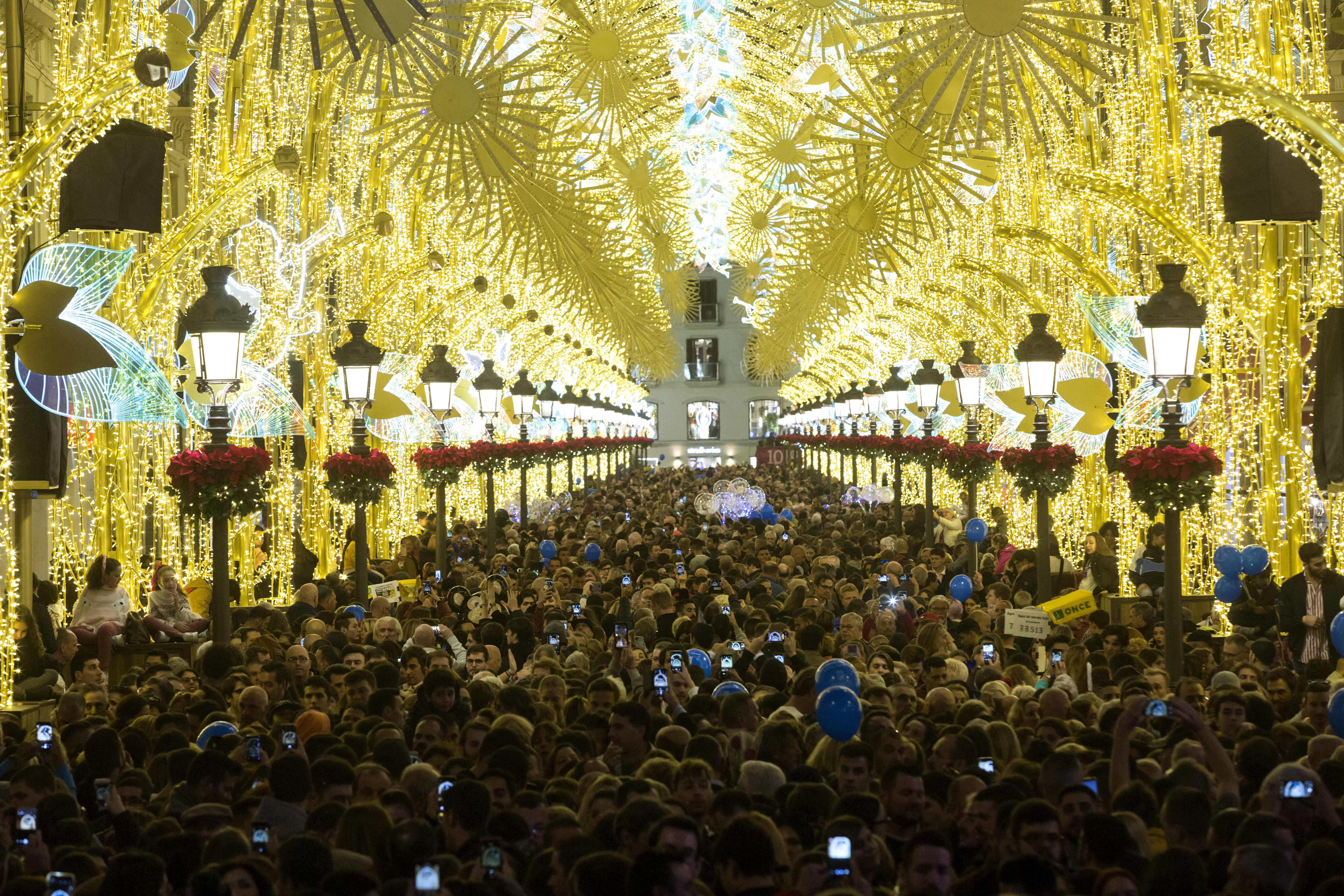 Gran multitud en calle decorada con luces de Navidad doradas. Farolas con poinsettias.