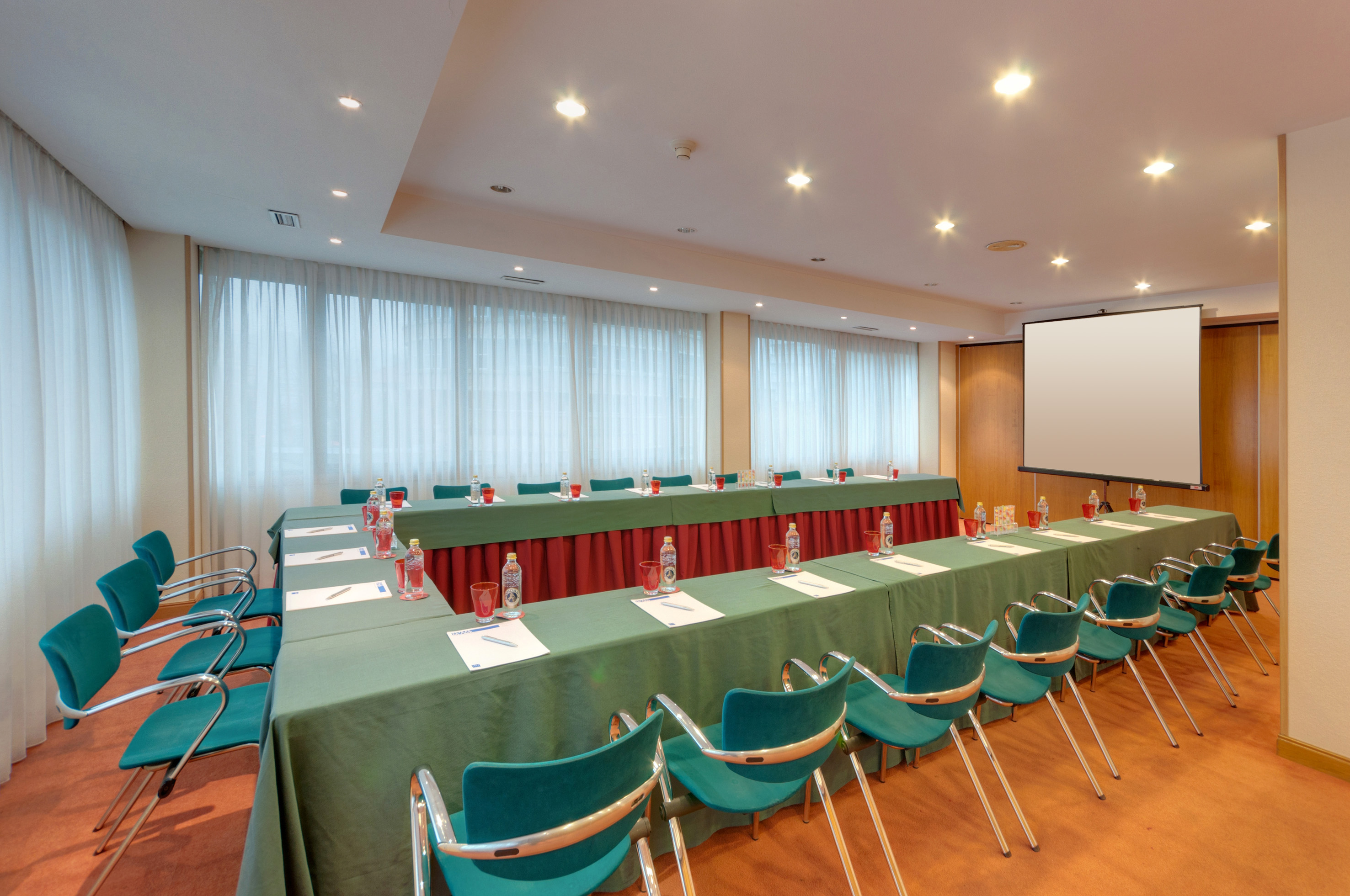 Conference room with U-shaped tables, teal chairs, water bottles, and a projector screen.