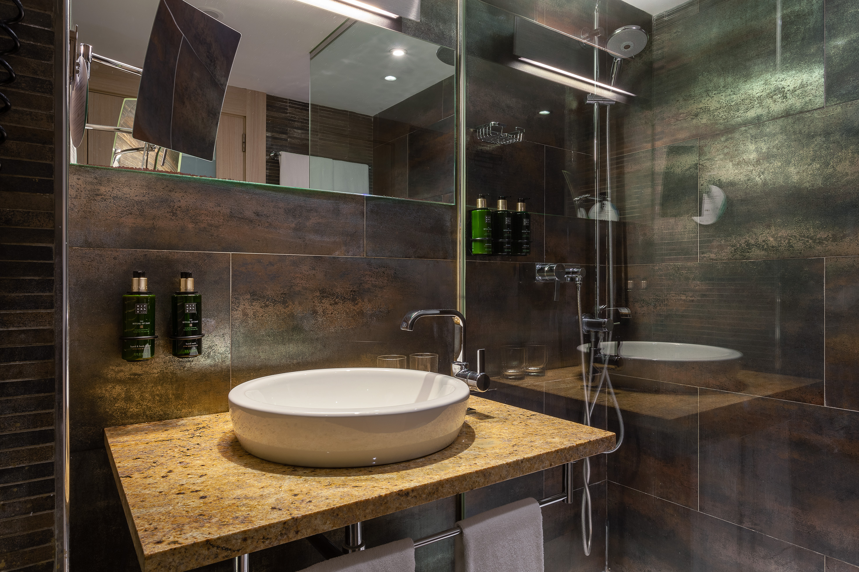 Modern hotel bathroom with granite counter, vessel sink, shower, and dark textured tiles.