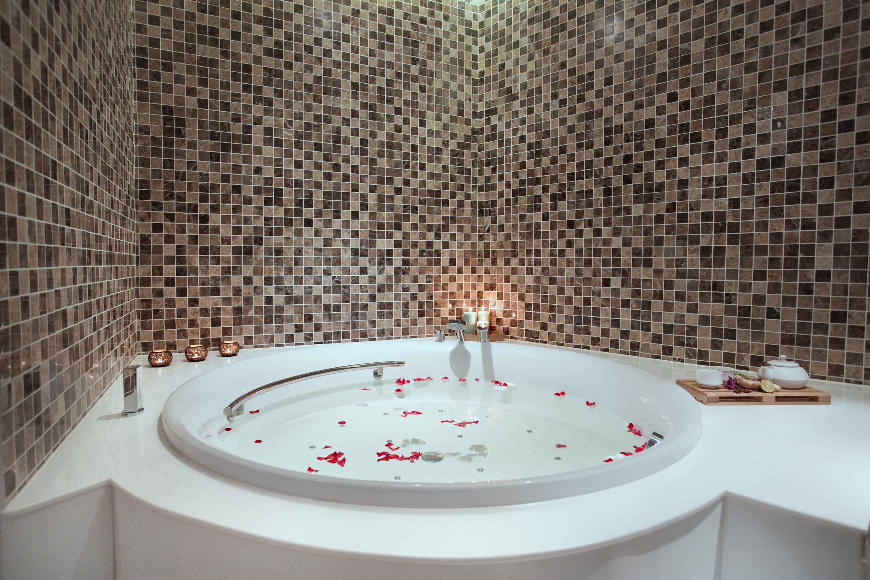 White spa bathtub with rose petals, lit candles, and mosaic wall tiles.