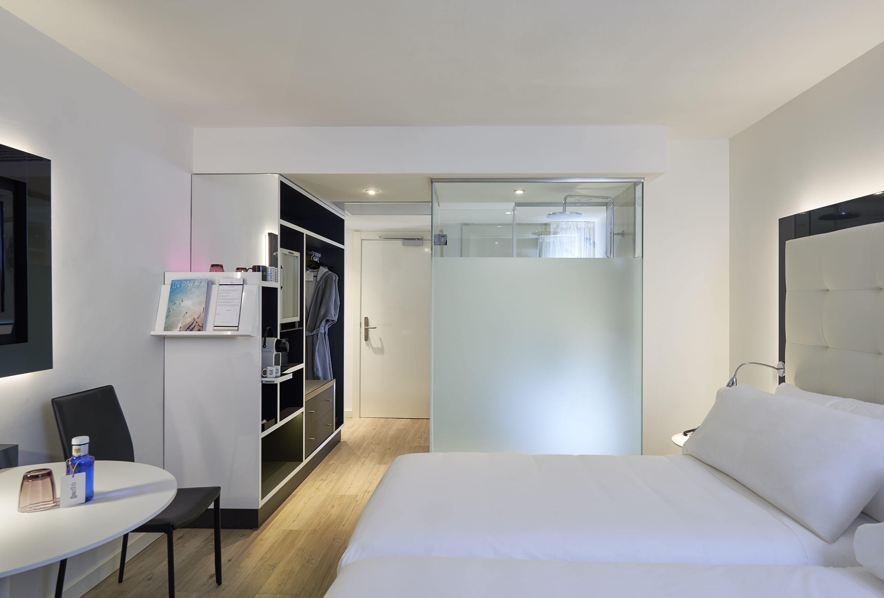 Modern hotel room with white bed, glass shower, and integrated storage.
