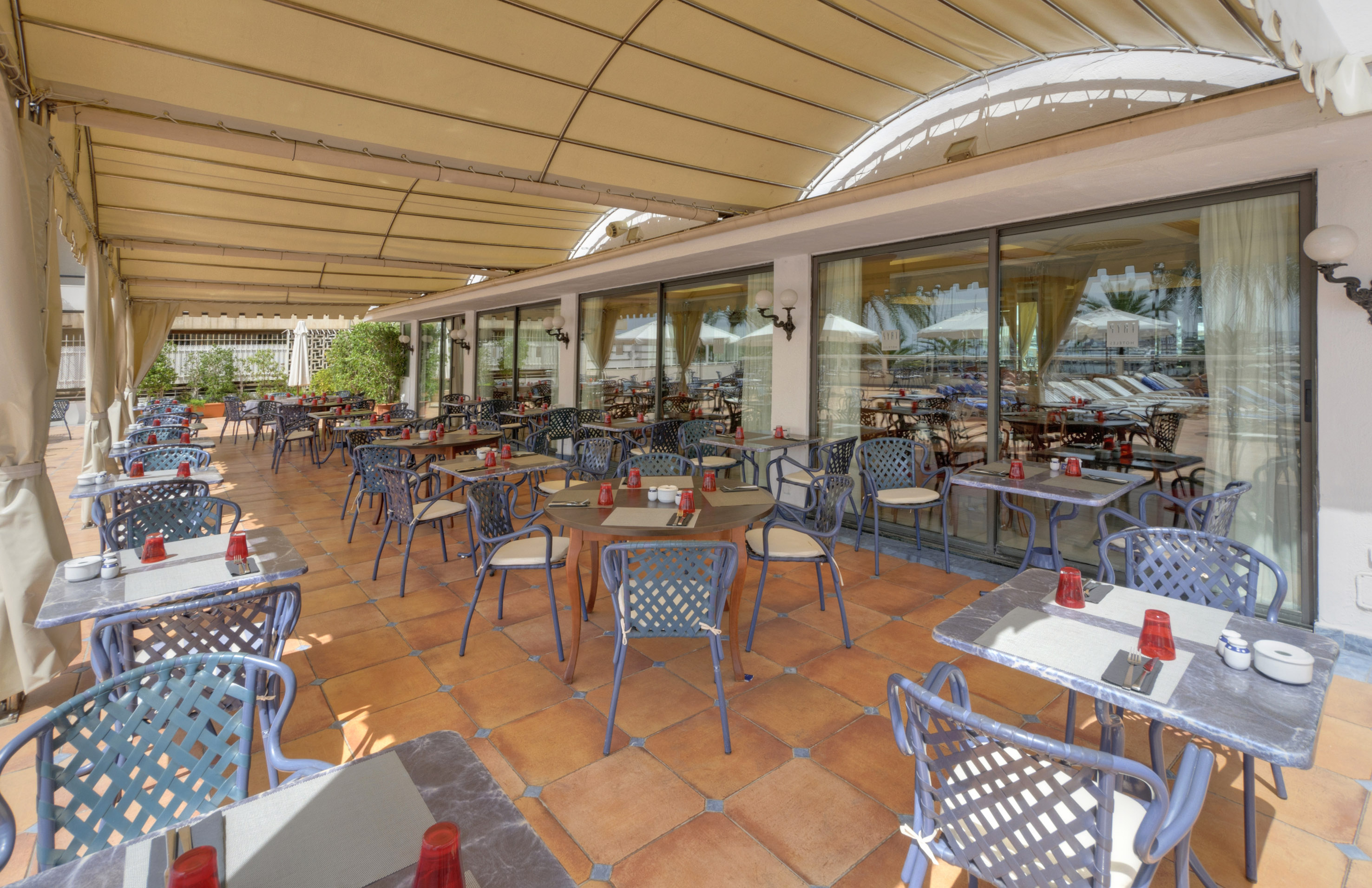 Outdoor dining patio with many tables, blue chairs, terracotta tiles, and awning.